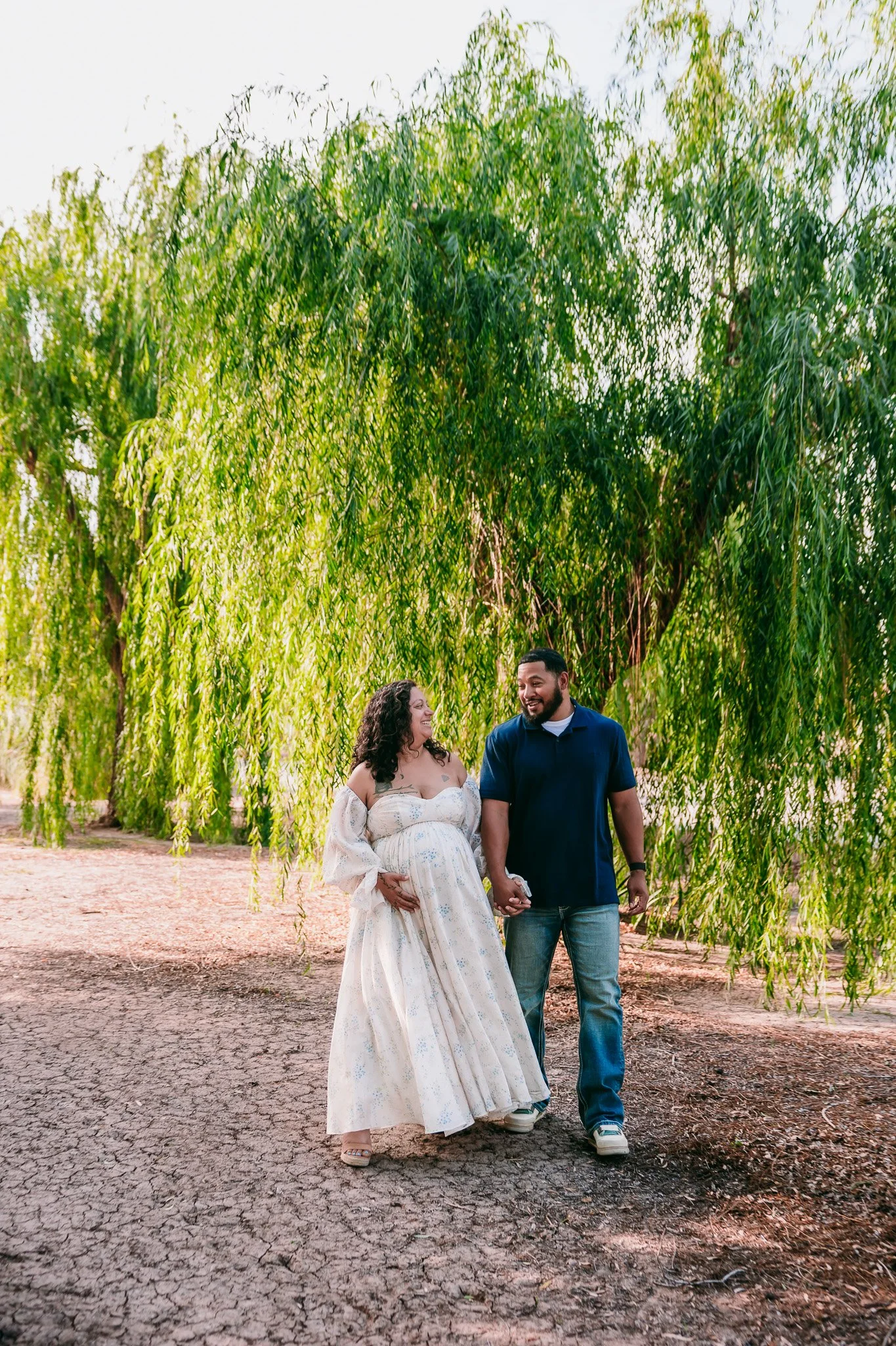 Expecting couple walking hand in hand beneath the willow trees during a maternity session in Las Cruces, New Mexico.