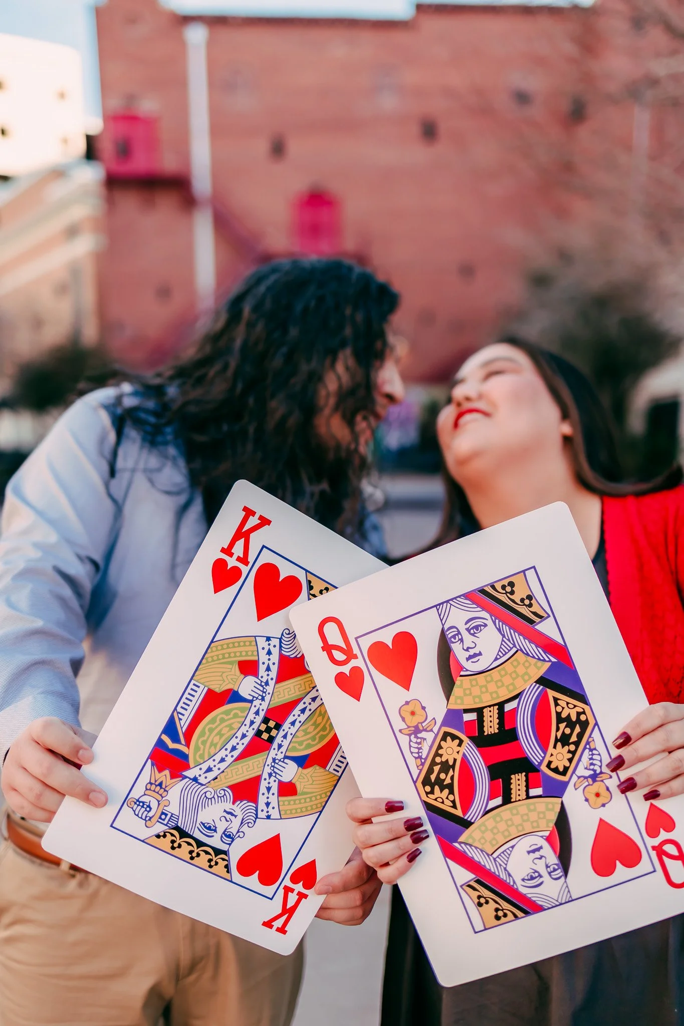 Partners sharing a kiss while holding oversized playing cards during a unique engagement session in downtown El Paso.