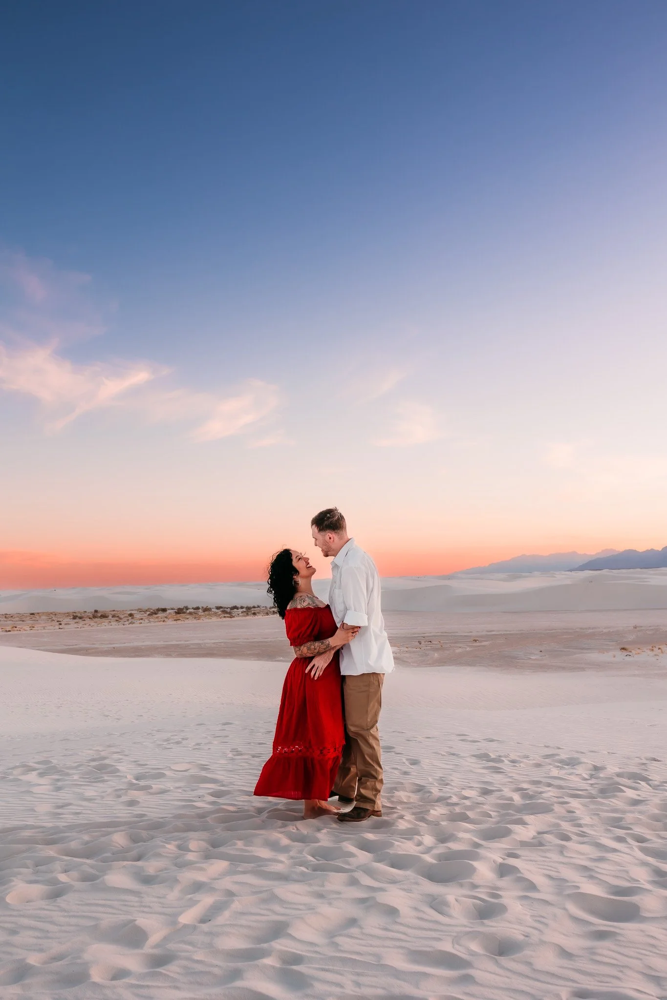 Engaged couple embracing beneath a colorful sunset sky at White Sands National Park during their engagement photography session.