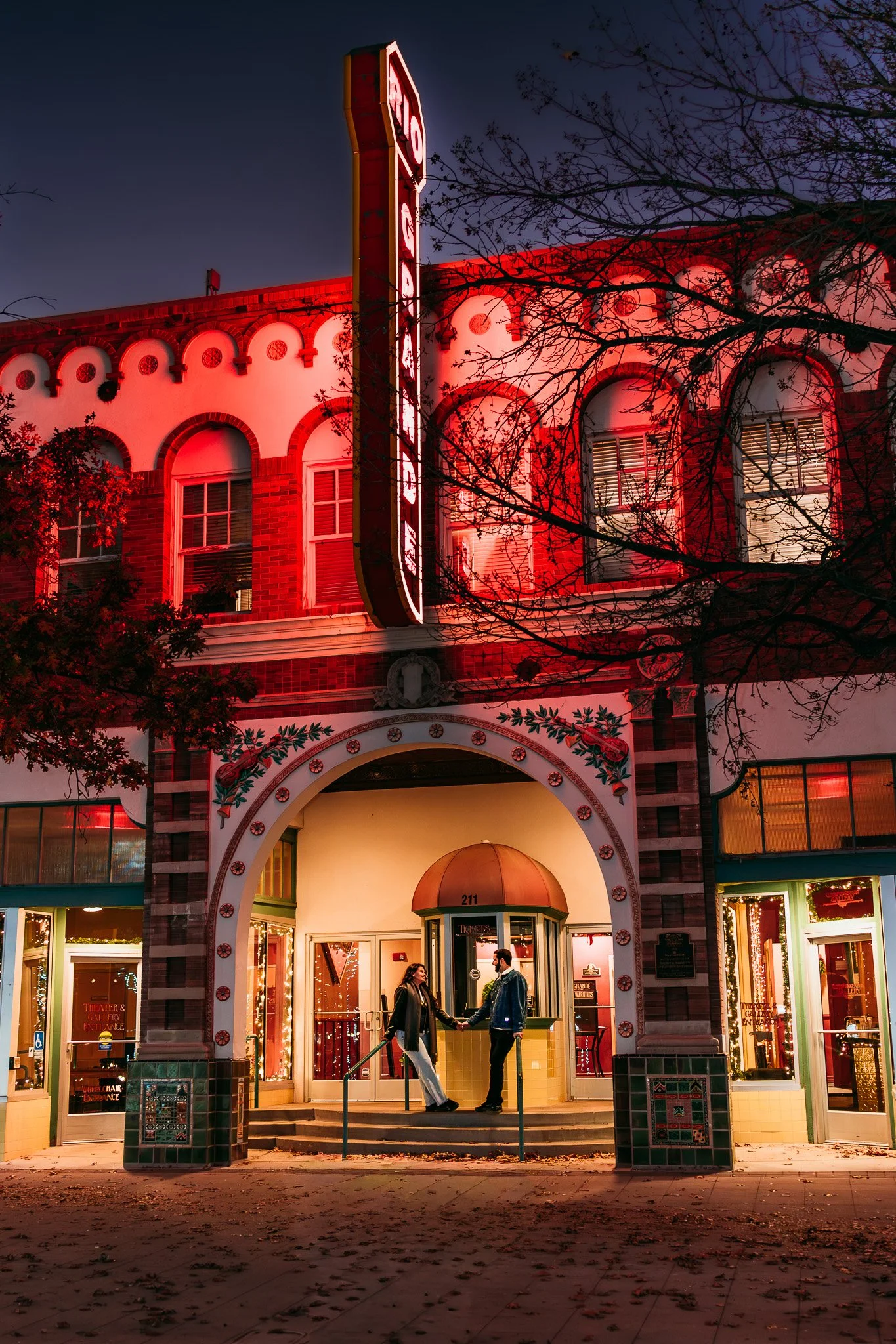 A neon-lit building with a couple shaking hands on the steps outside during the evening. The building has arched windows and vibrant red, pink, and yellow lighting, with some tree branches visible in the foreground.