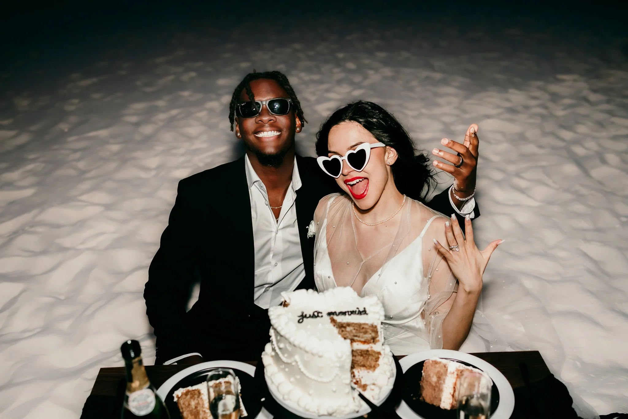 Newly married couple wearing heart-shaped sunglasses while celebrating with cake and champagne after their White Sands National Park elopement.