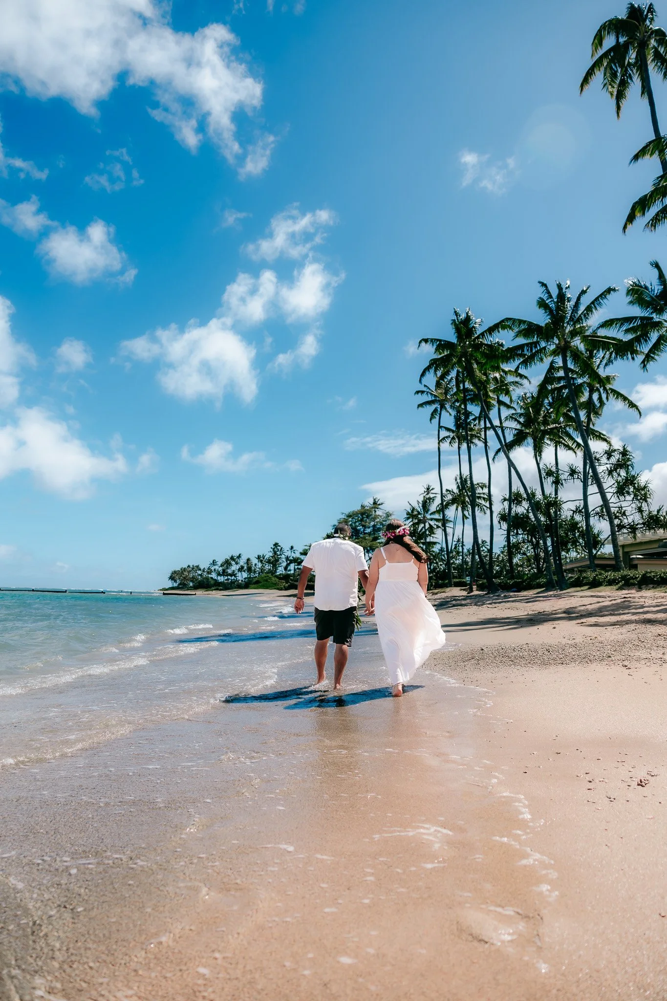 Bride and groom walking hand in hand along the beach shore during their destination wedding on Oahu, Hawaii.