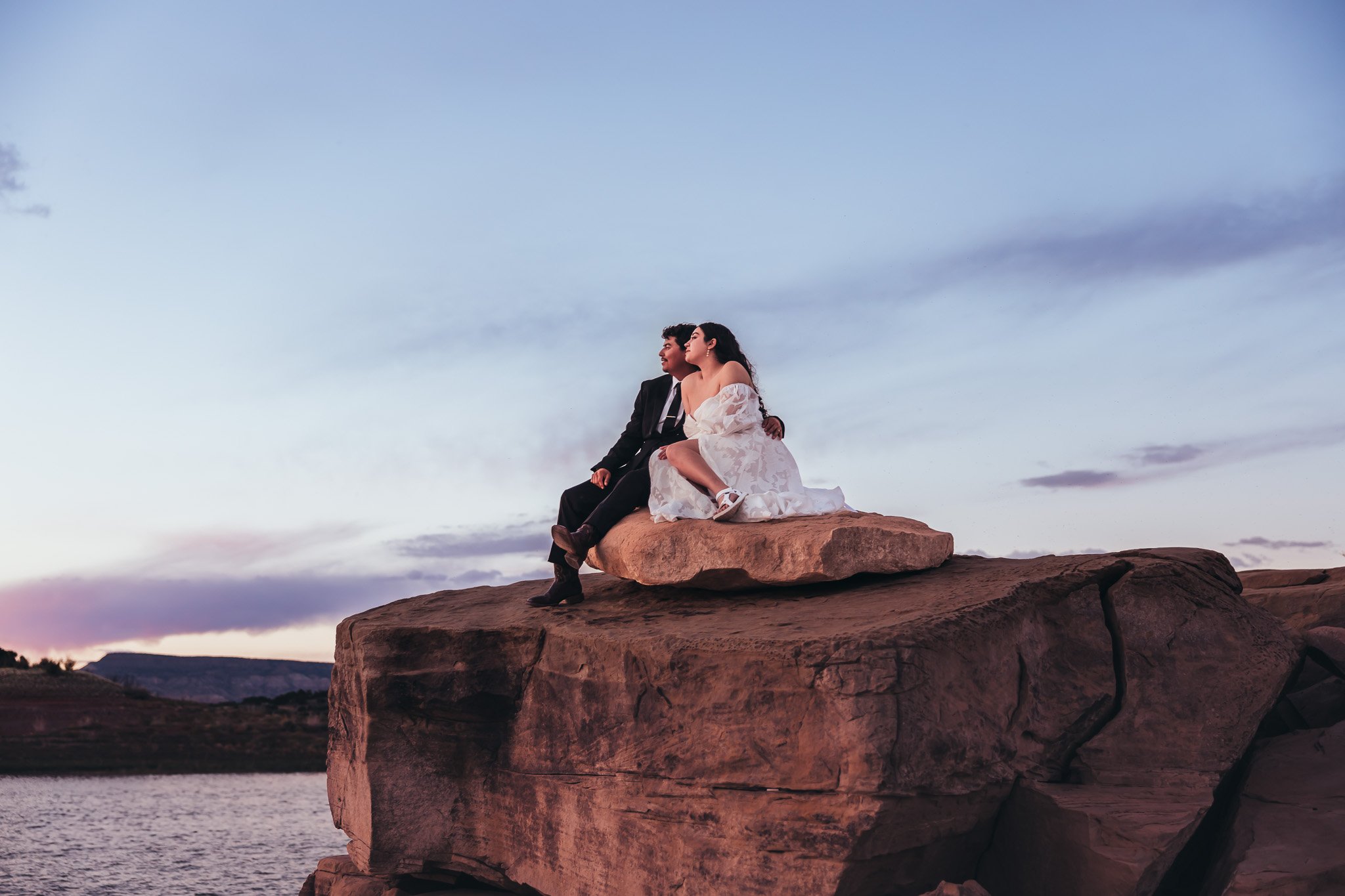 Newly married couple sitting together on dramatic rock formations overlooking Abiquiu Lake during their New Mexico elopement at sunset.
