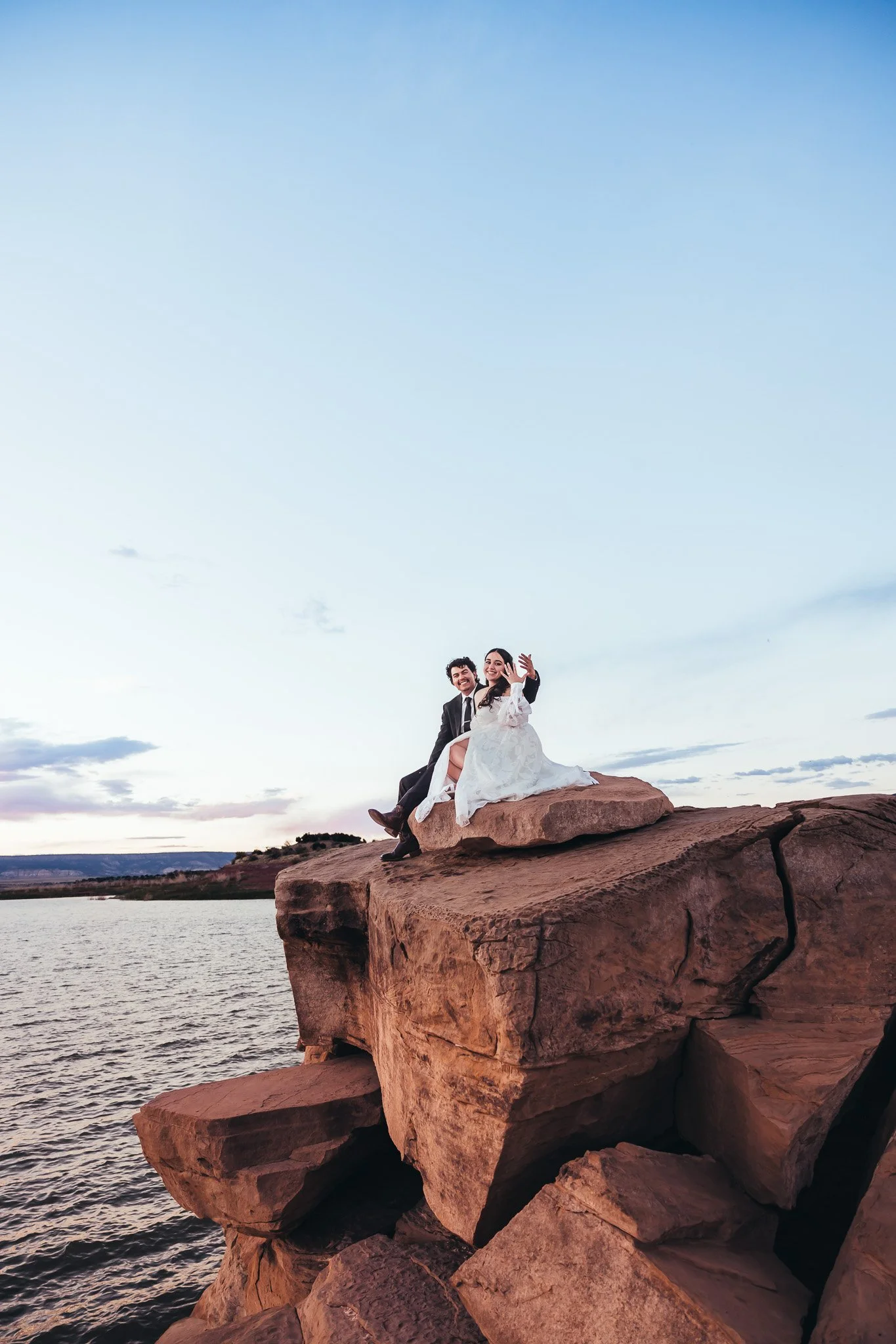Newly married couple sitting together on dramatic rock formations overlooking Abiquiu Lake during their New Mexico showing off their rings at elopement at sunset.