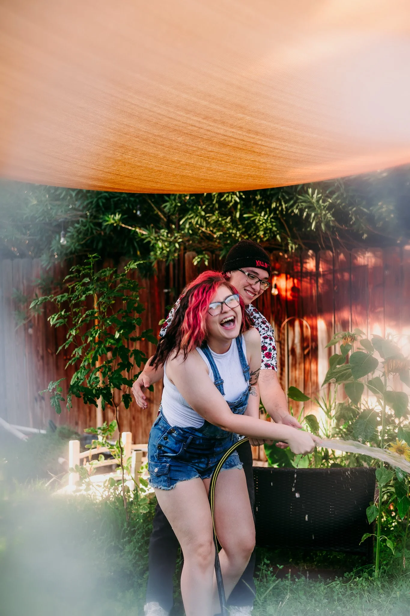 Couple laughing together while playing with a garden sprinkler during a playful backyard couples session at their home in Las Cruces, New Mexico.