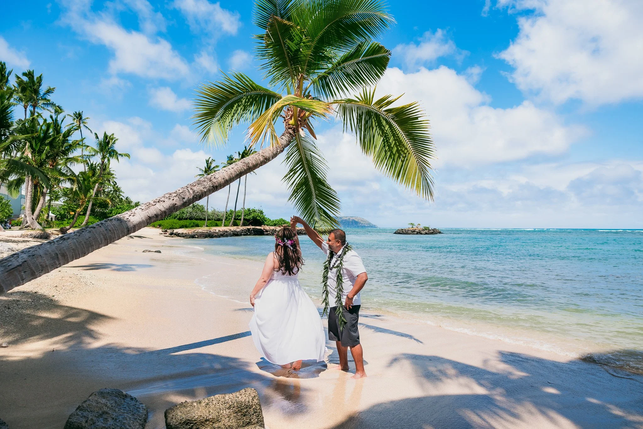 Newlyweds dancing under an iconic palm tree on the beach during their destination elopement on Oahu, Hawaii.