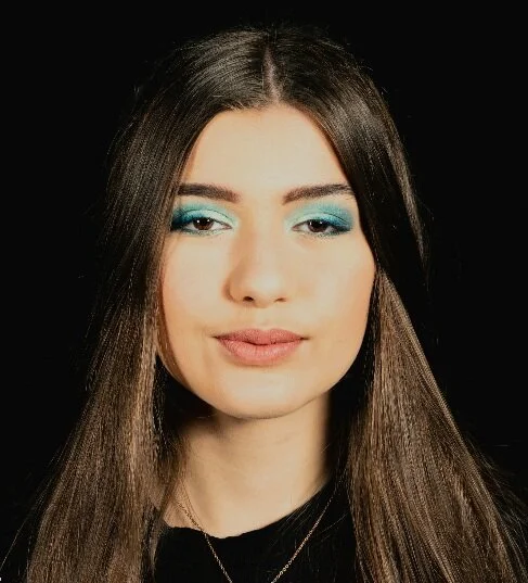 Porträt einer jungen Frau mit langen braunen Haaren, blauer Make-up auf den Augen, vor schwarzem Hintergrund.