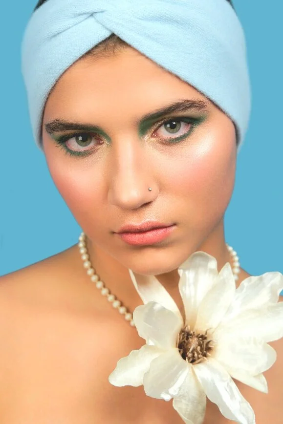 Junge Frau mit blauer Kopftuch, blauer Hintergrund, Perlenkette, hält eine große weiße Blume, trägt Make-up und hat einen Nasenpiercing.