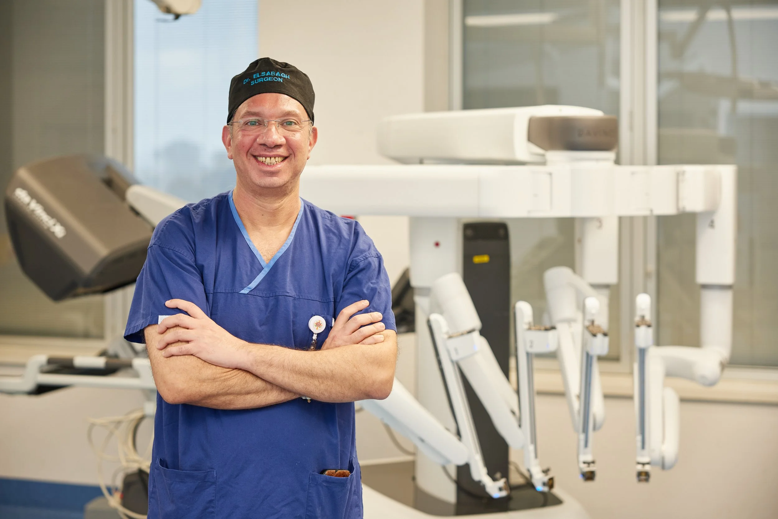 A smiling male doctor in blue scrubs and a black cap standing with arms crossed in a hospital room with medical equipment.