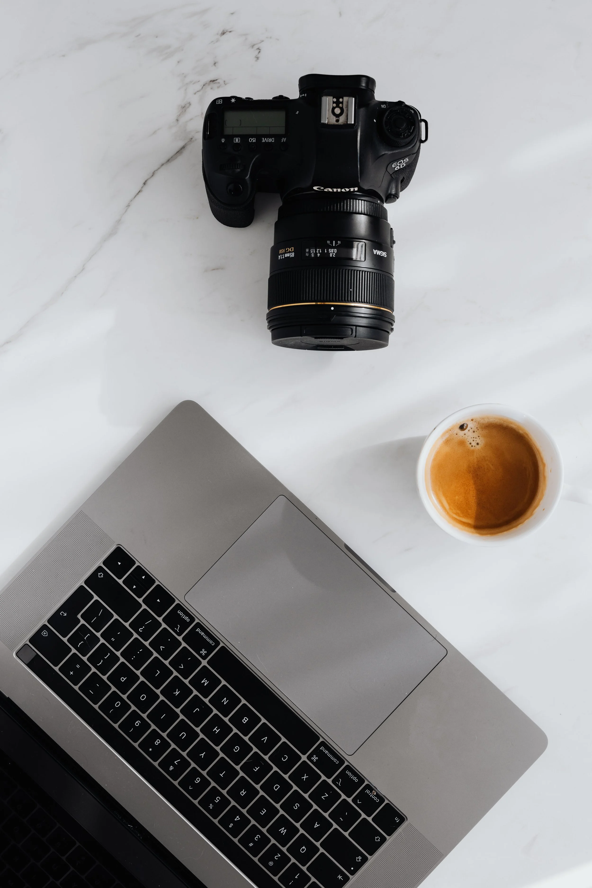 A flat lay of a workspace with a silver laptop, a black DSLR camera, a cup of coffee, all on a white marble surface.