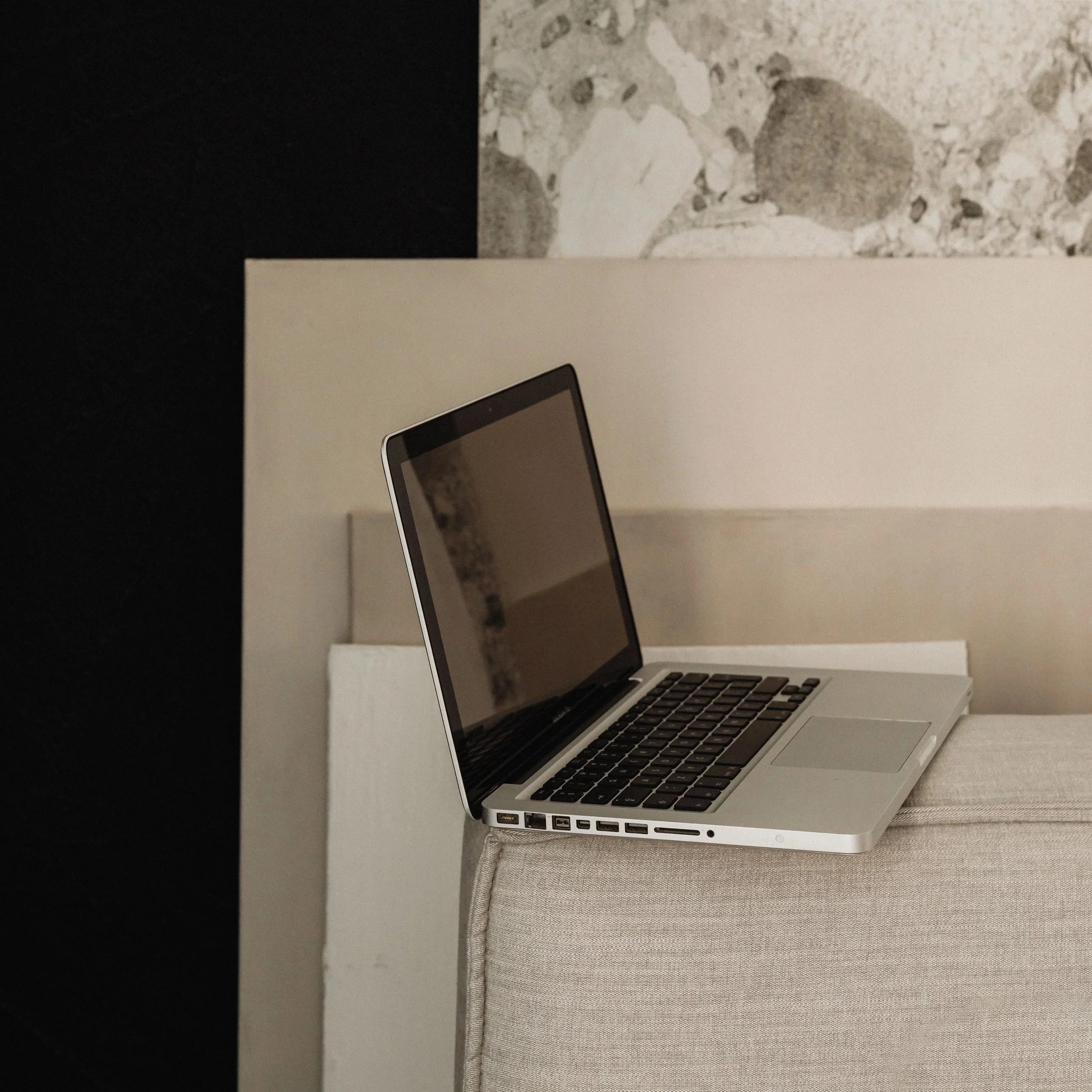 Silver laptop with black keyboard resting on beige upholstered furniture with a textured pillow, against a background of black, white, and gray decor.