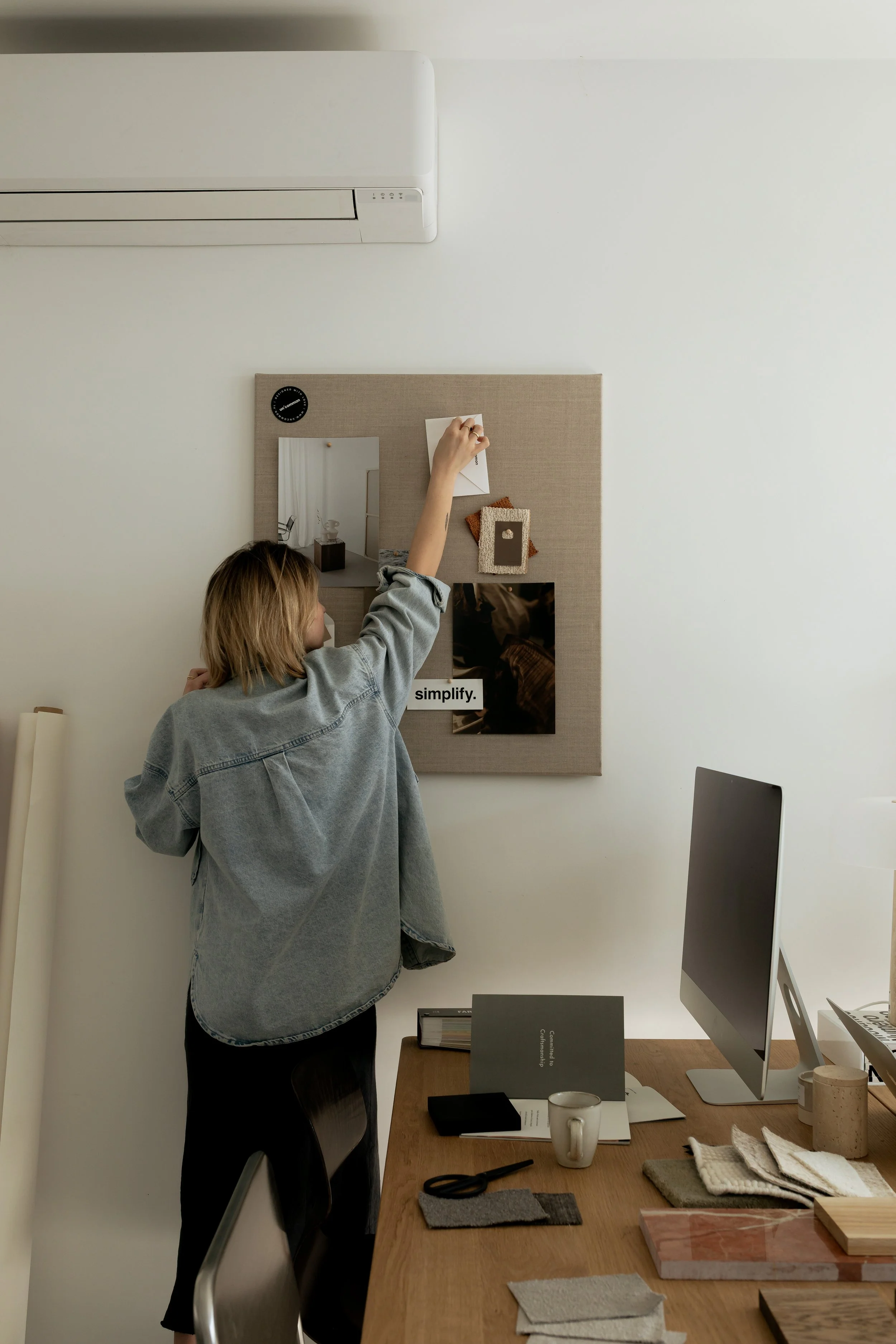 A woman arranging notes or pictures on a corkboard in a modern workspace with a computer, mug, and design samples on the desk.