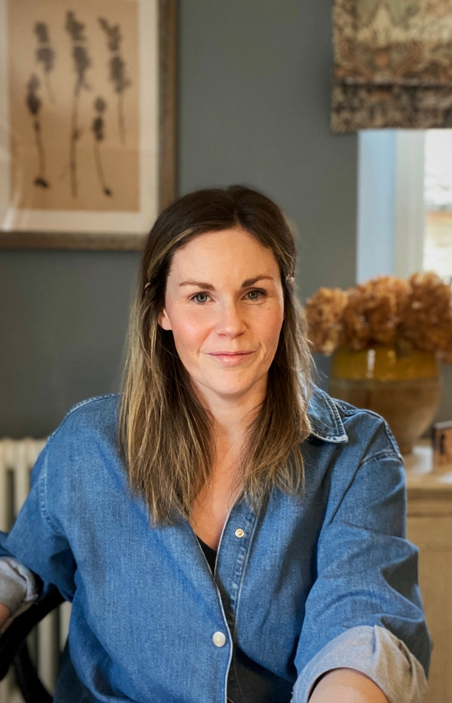 A woman with long brown hair and blue eyes wearing a denim jacket, sitting at a table in a cozy room with blue walls, artwork on the wall, and a flower arrangement in the background.