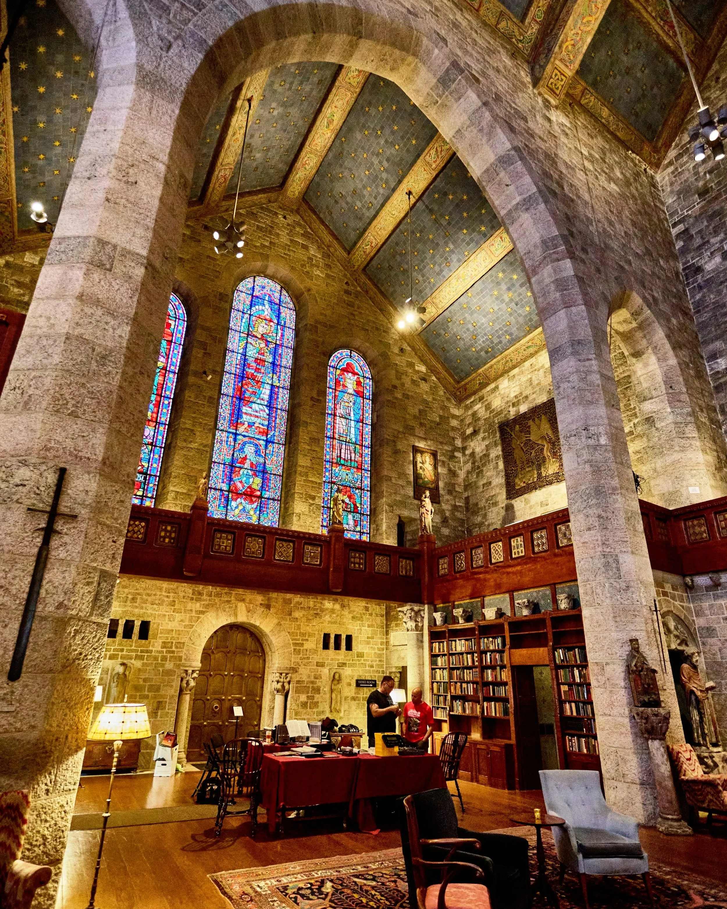 Interior of a historic church or cathedral with stained glass windows, stone walls, and wooden furnishings, including tables and sculptures.