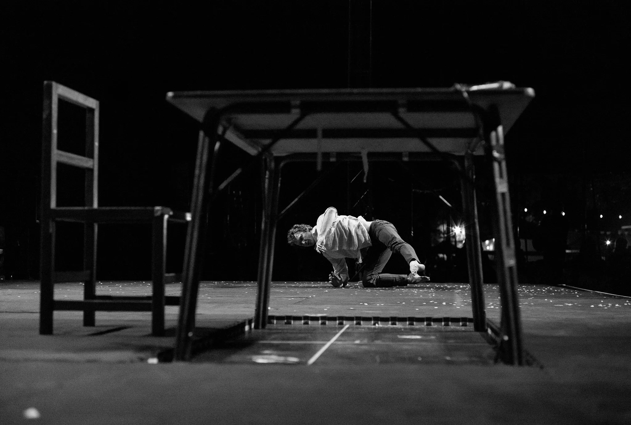 Acrobat Lewie West is crouched on the floor of a dimly lit stage, reaching towards the camera, peering underneath a table with an empty chair in the foreground