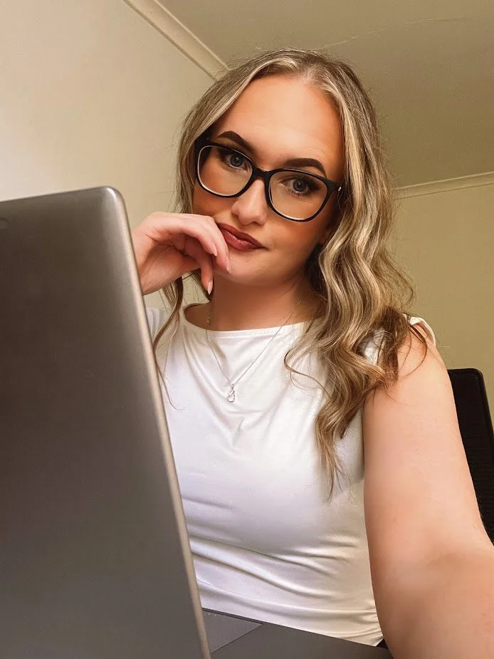 A woman with wavy blonde hair, wearing glasses and a white sleeveless top, sitting at a desk with a silver laptop, looking at the camera with a thoughtful expression.