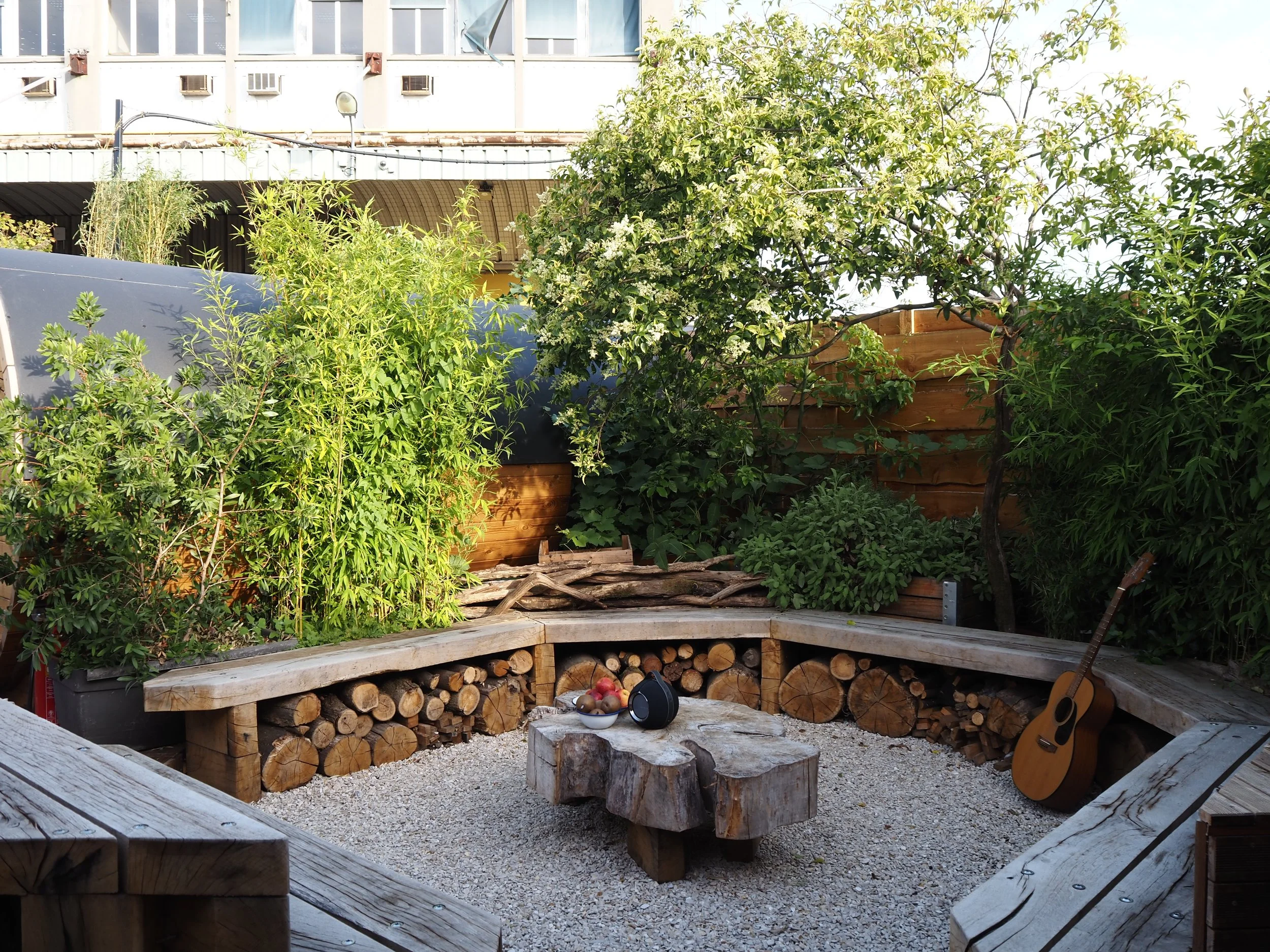 People relaxing and socializing on an outdoor rooftop terrace, with some sitting on a bench, others standing, and a few inside a small wooden sauna or steam room. The area has potted plants, shoes outside, and a sign reading 'Silent Sauna'.