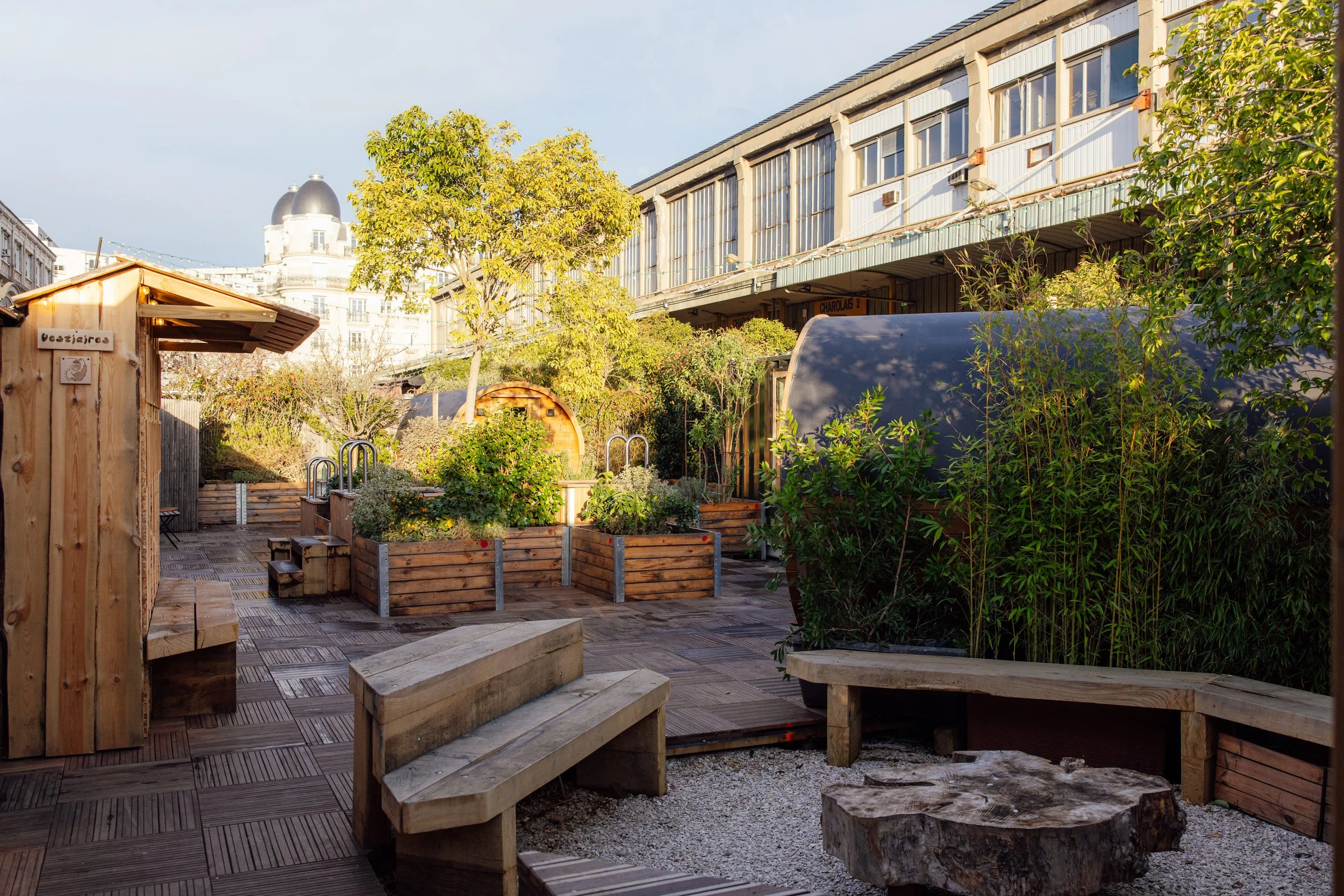 People relaxing and socializing on an outdoor rooftop terrace, with some sitting on a bench, others standing, and a few inside a small wooden sauna or steam room. The area has potted plants, shoes outside, and a sign reading 'Silent Sauna'.