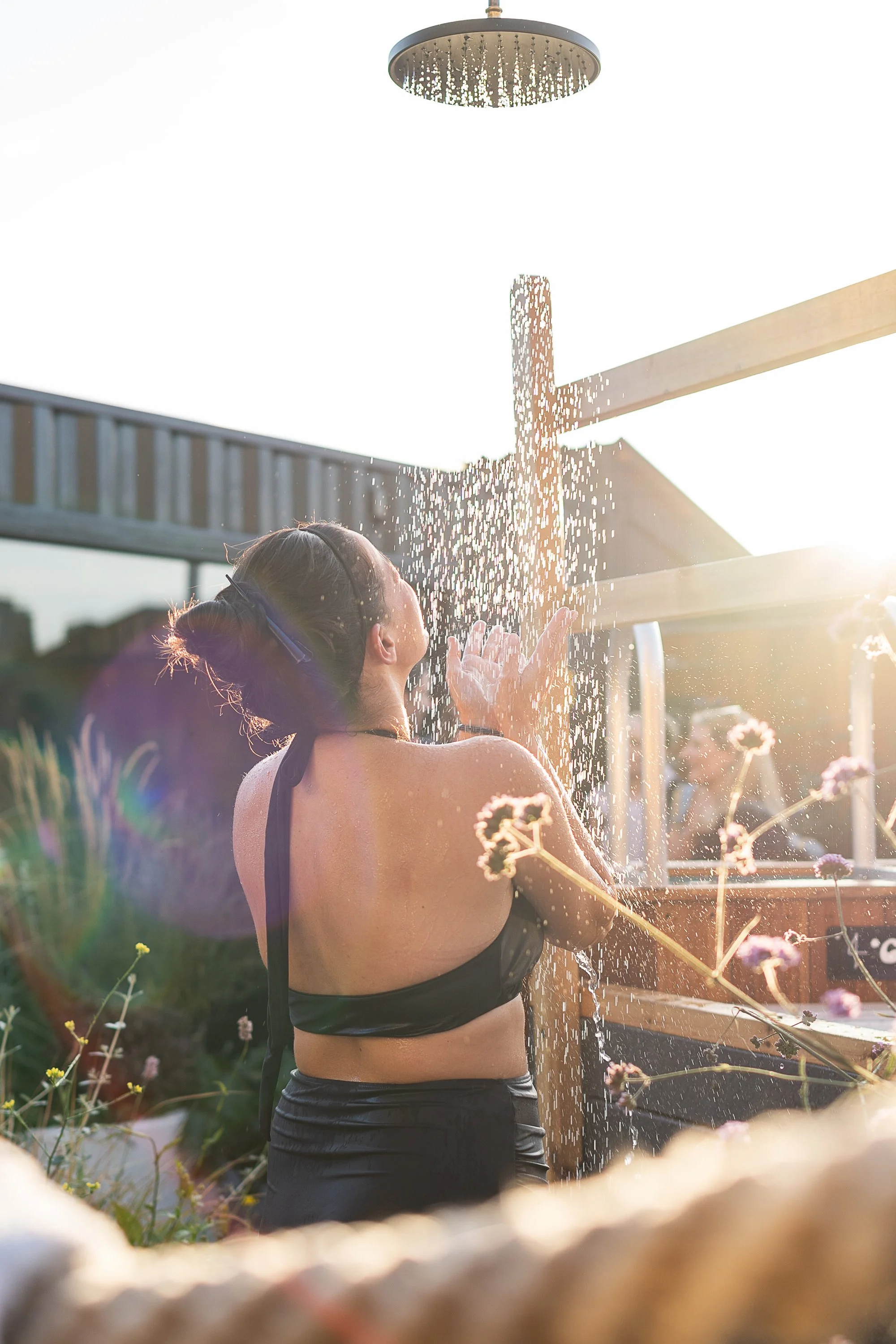 A woman in a black swimsuit enjoying an outdoor shower with sunlight shining behind, surrounded by flowers.