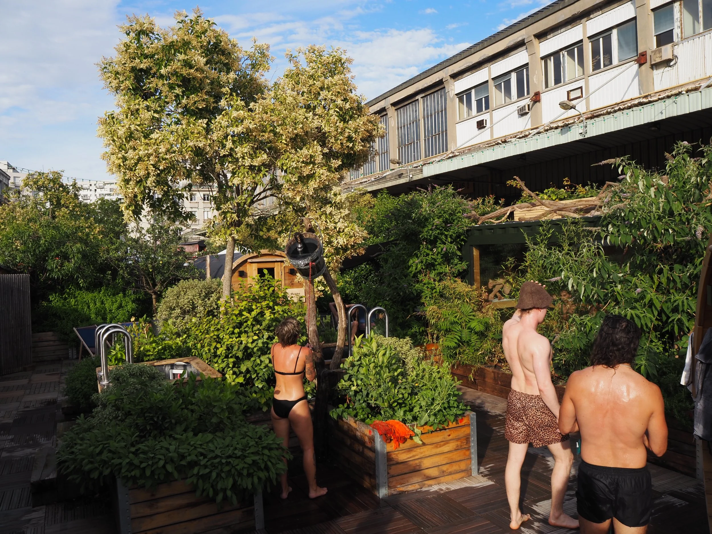A person relaxing in a bathtub outdoors surrounded by plants and wooden structures, with a wooden barrel, a small wooden building, lush greenery, and a cityscape background.