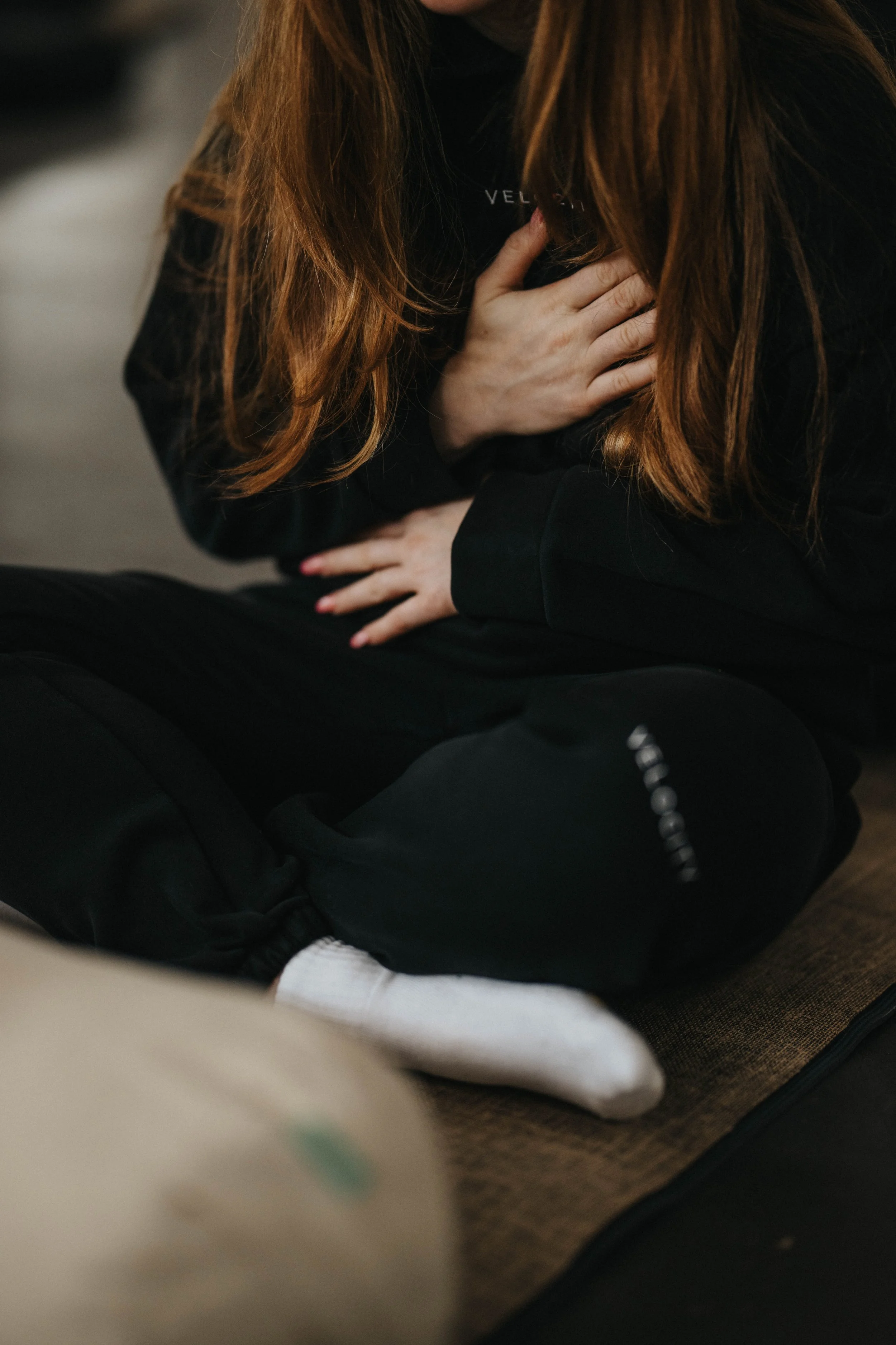 A person sitting on a carpeted floor, clutching their chest with one hand and holding their stomach with the other, experiencing discomfort or pain.