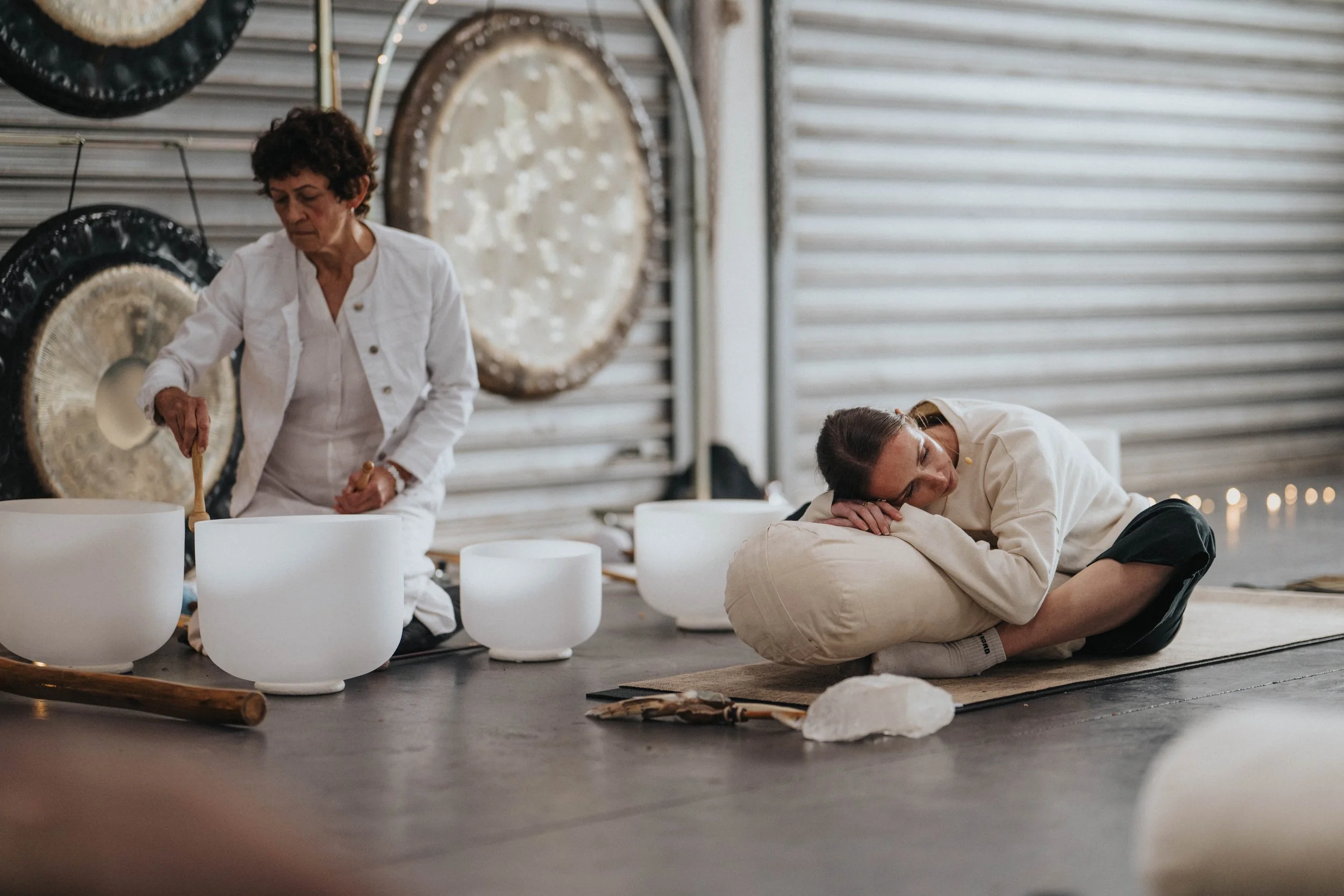 A woman playing crystal singing bowls while a young man sleeps on a cushion on the floor in a yoga studio or wellness space with large gongs hanging on the wall.