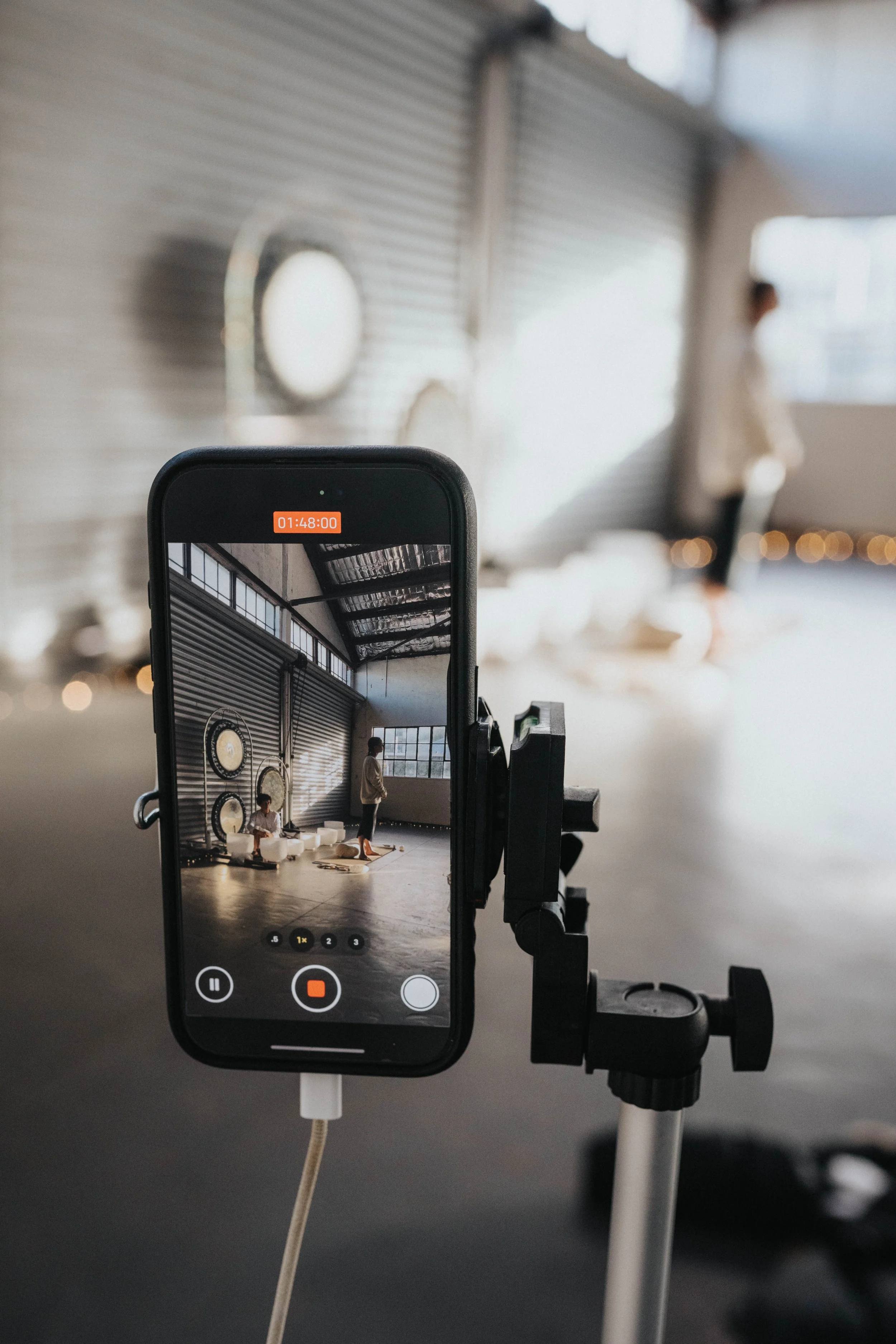 A smartphone mounted on a tripod recording a video or taking a photo of two people in a spacious industrial-style room with large windows and corrugated metal walls.