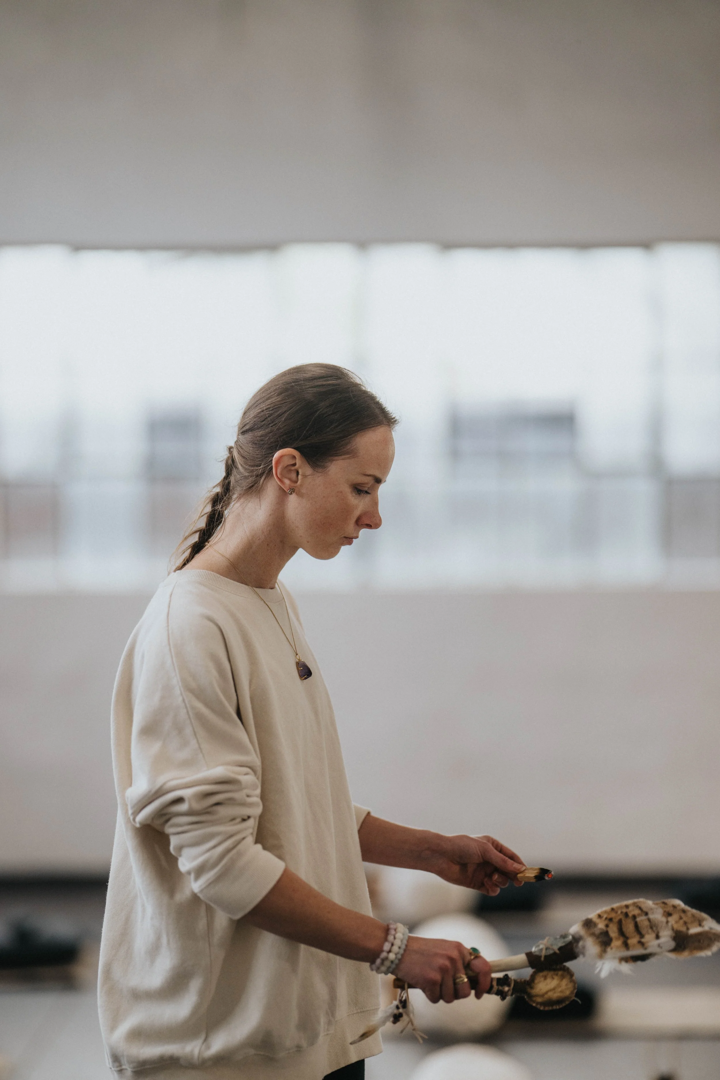 A woman with a braid and light beige sweater holding burnt offering sticks, standing in a room with large windows in the background.