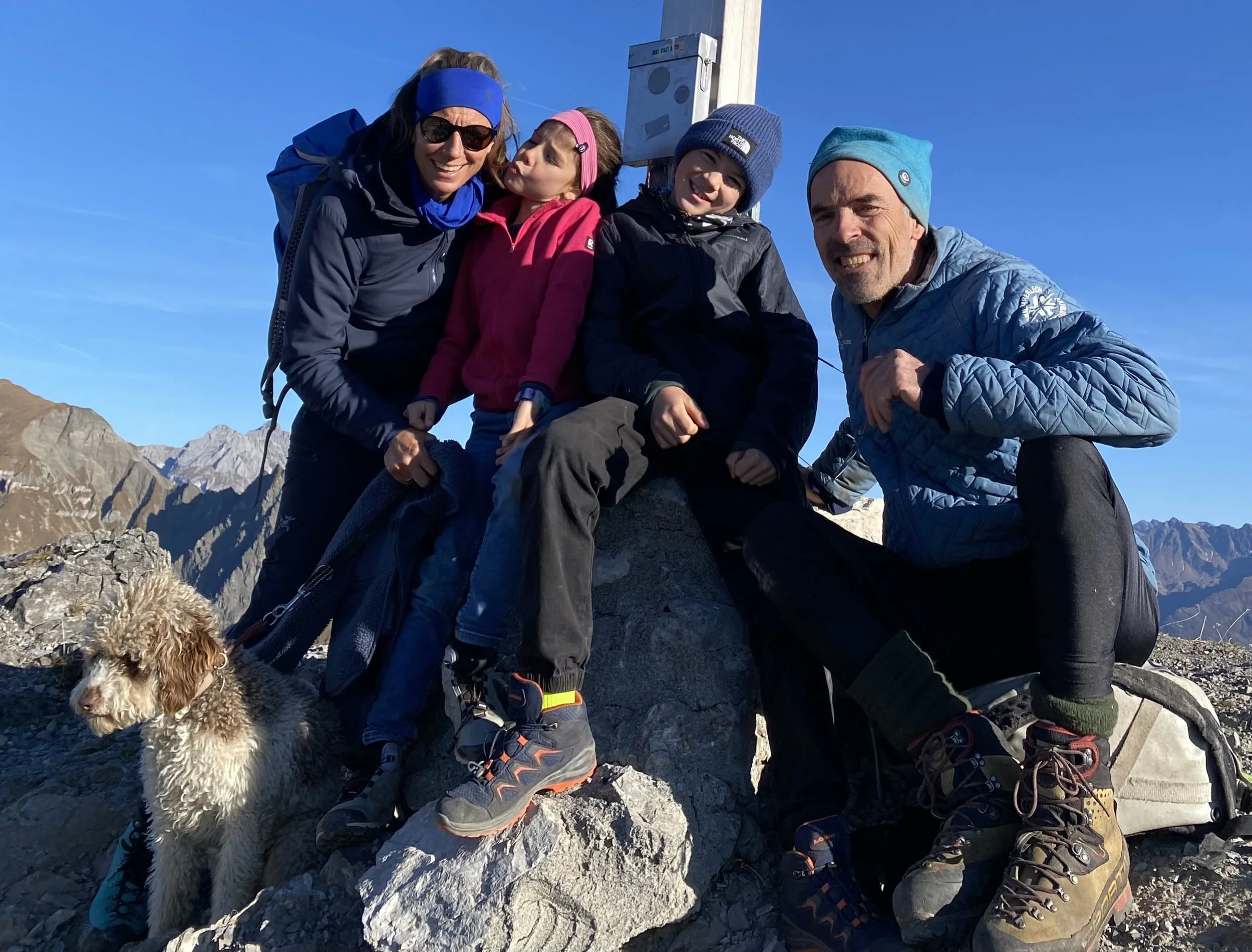 Familie mit zwei Kindern und einem Hund auf einem Berggipfel, umgeben von Bergen, mit blauer Kleidung und Outdoor-Ausrüstung.