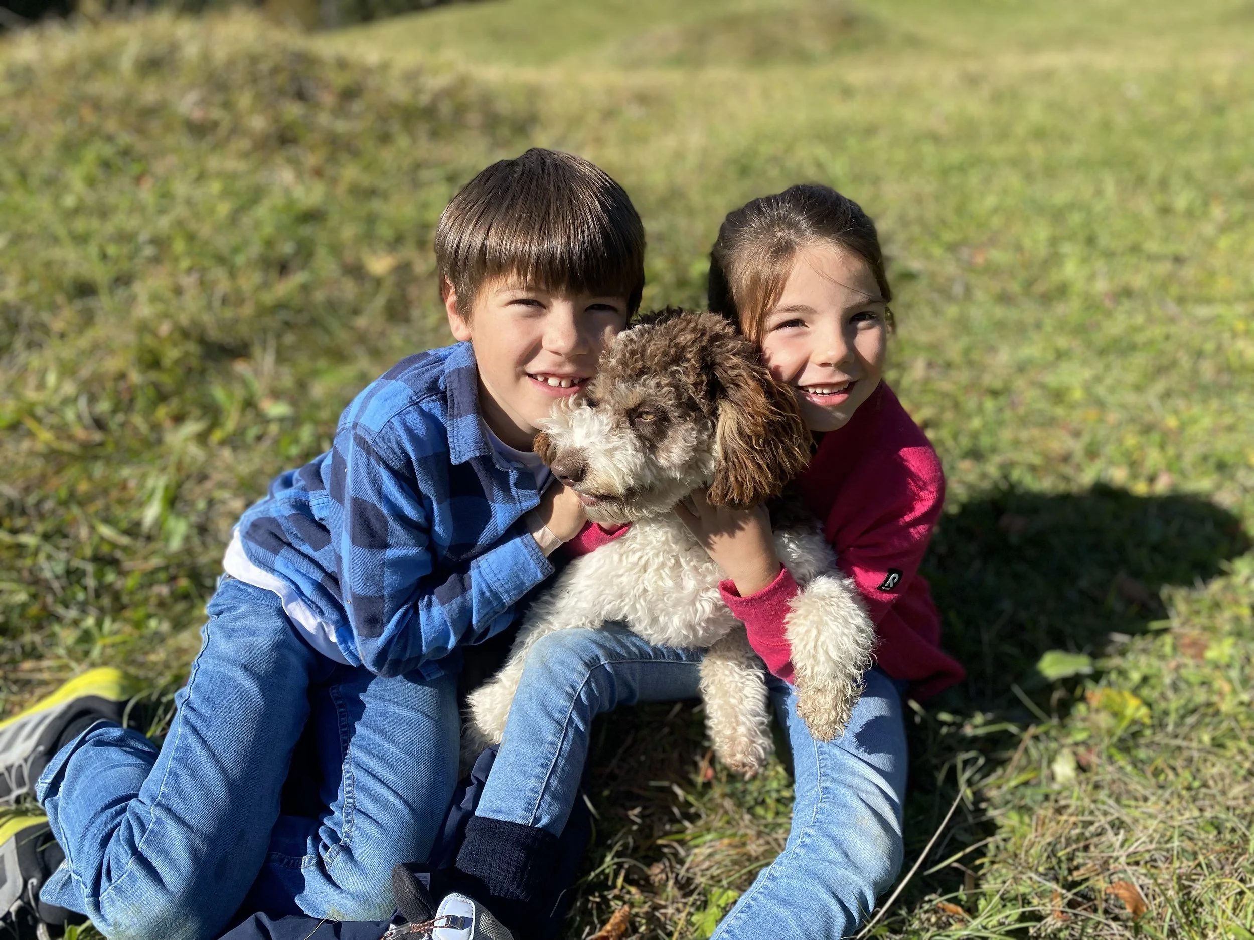 Zwei lachende Kinder, ein Junge in blauer Jacke und eine Mädchen in roter Jacke, sitzen im Freien auf dem Gras, während sie ein braun-weißen Hund halten.