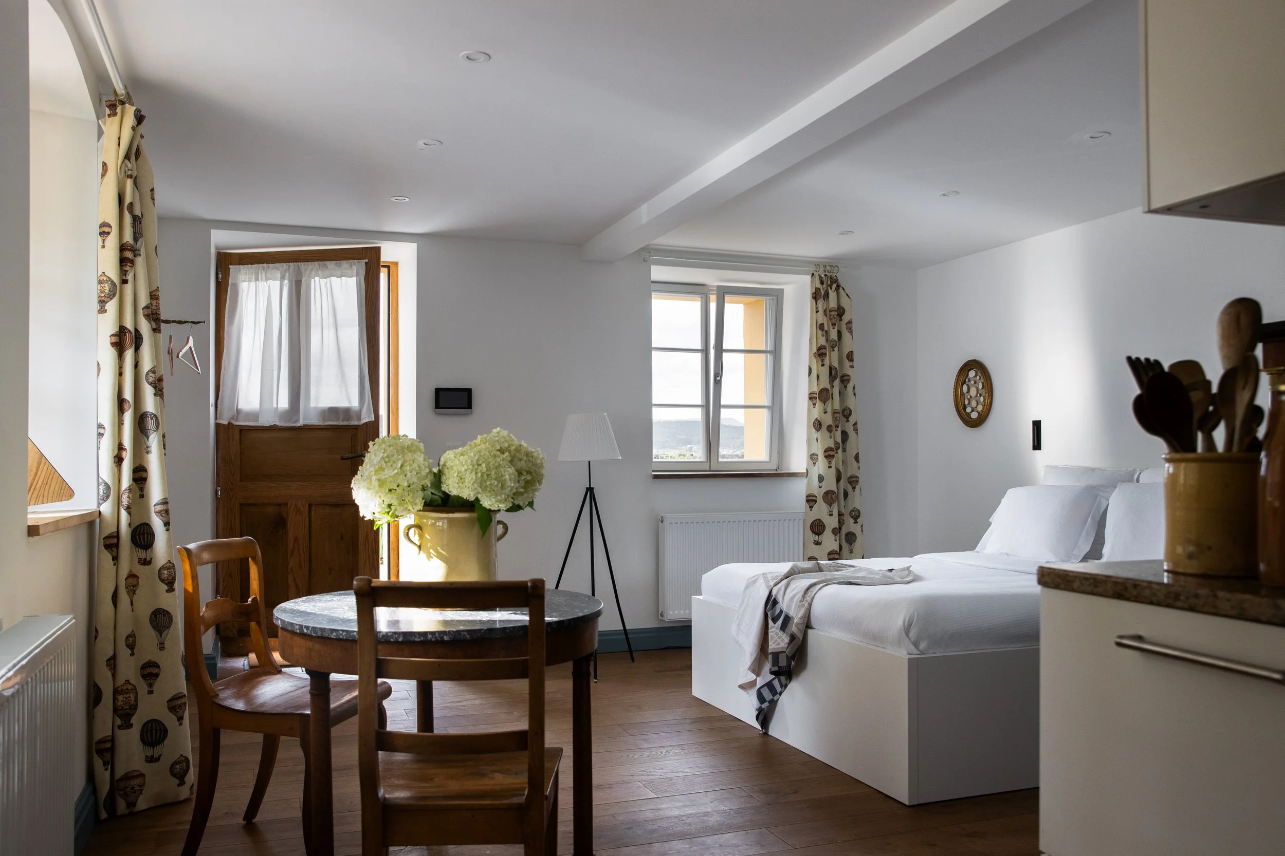 Intérieur d'une chambre lumineuse au Château de Marnand avec lit, fenêtres, rideaux à motifs de Montgolfières et décoration chaleureuse.