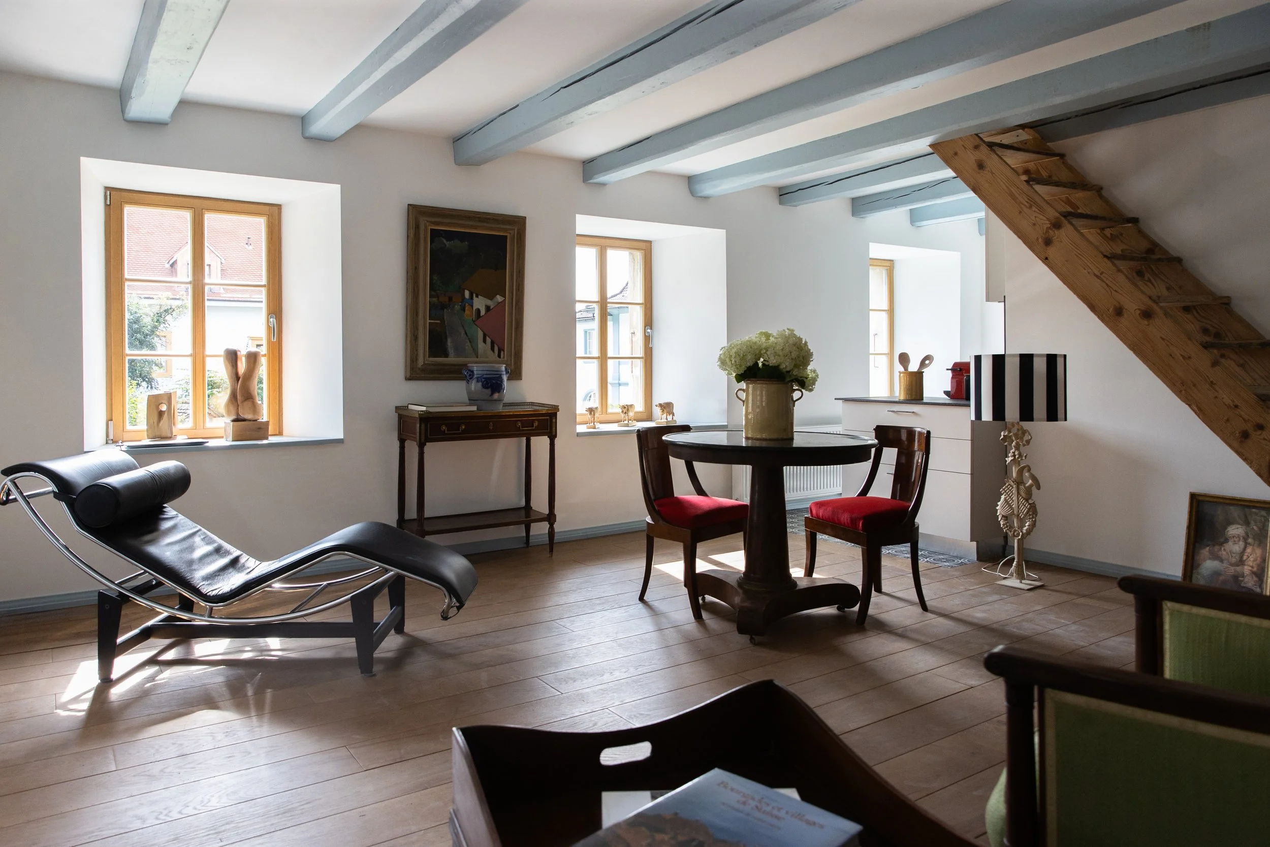 Intérieur d'une pièce avec fenêtres en bois, meubles anciens, et une escalier en bois. Décorations artistiques sur les rebords des fenêtres et un tableau sur le mur.