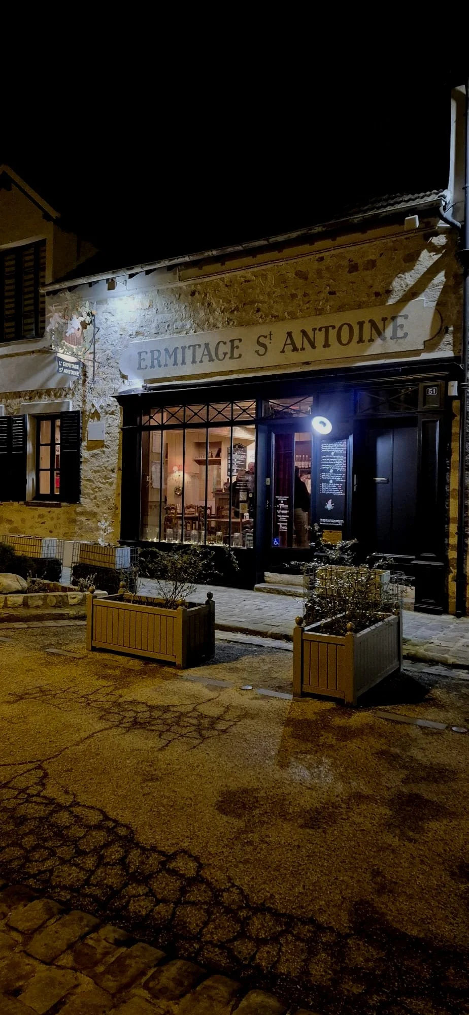 Une façade d'une boutique nommée "Ermitage St Antoine" éclairée la nuit, avec des fenêtres en verre et une porte noire, entourée de pots de fleurs en bois, sur une rue pavée.
