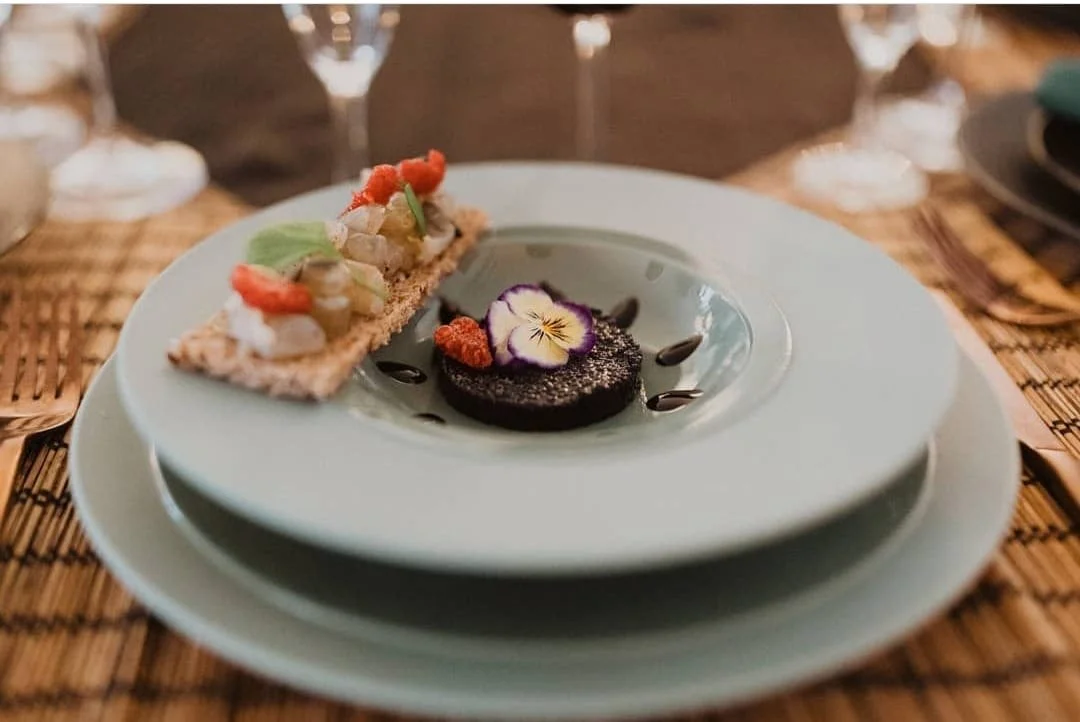 Assiette avec un dessert à gauche, probablement une tartelette tartare de fruits ou de poisson, et à droite, un gâteau au chocolat garni d'une fleur comestible violette et de décorations de sauce chocolat, sur une table en bois avec des verres à vin en arrière-plan.