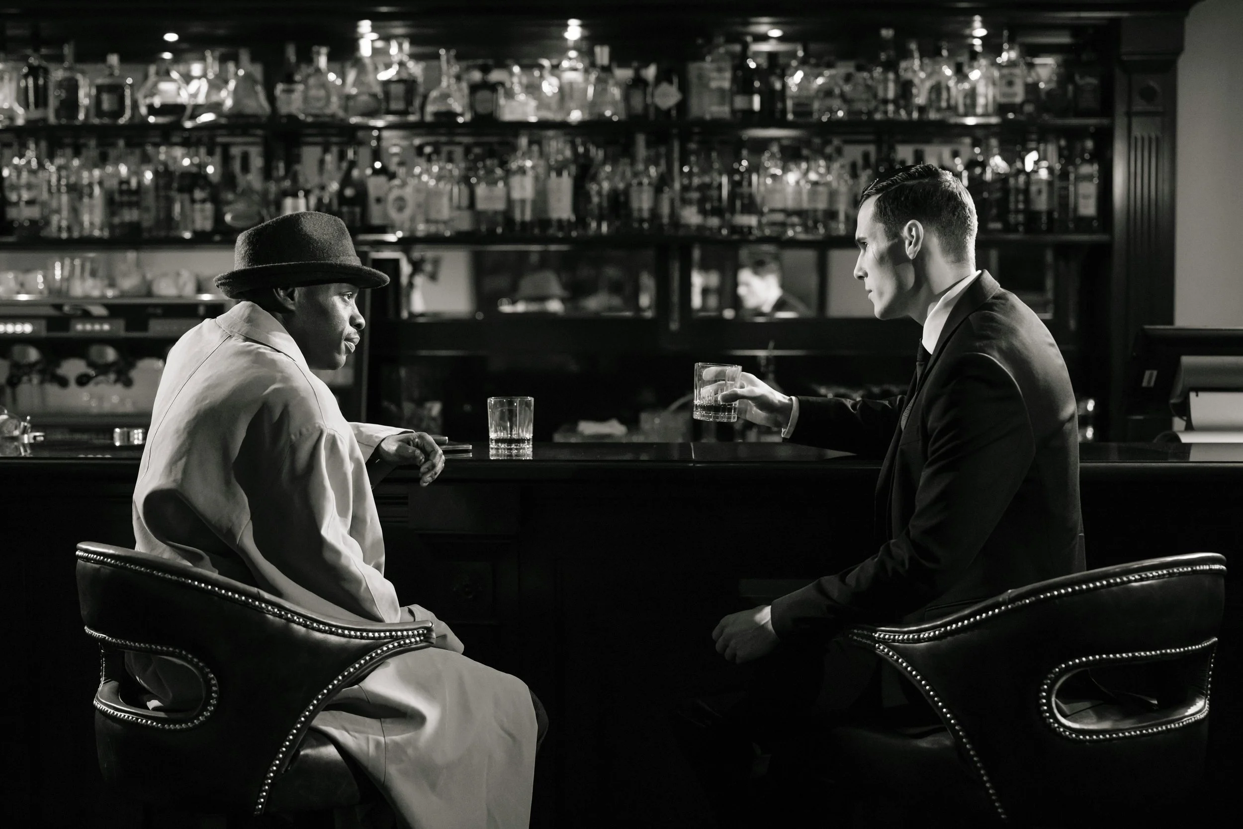 Deux hommes assis dans un bar, en train de boire un verre, en noir et blanc. L'un porte un chapeau et une longue veste, l'autre est en costume.