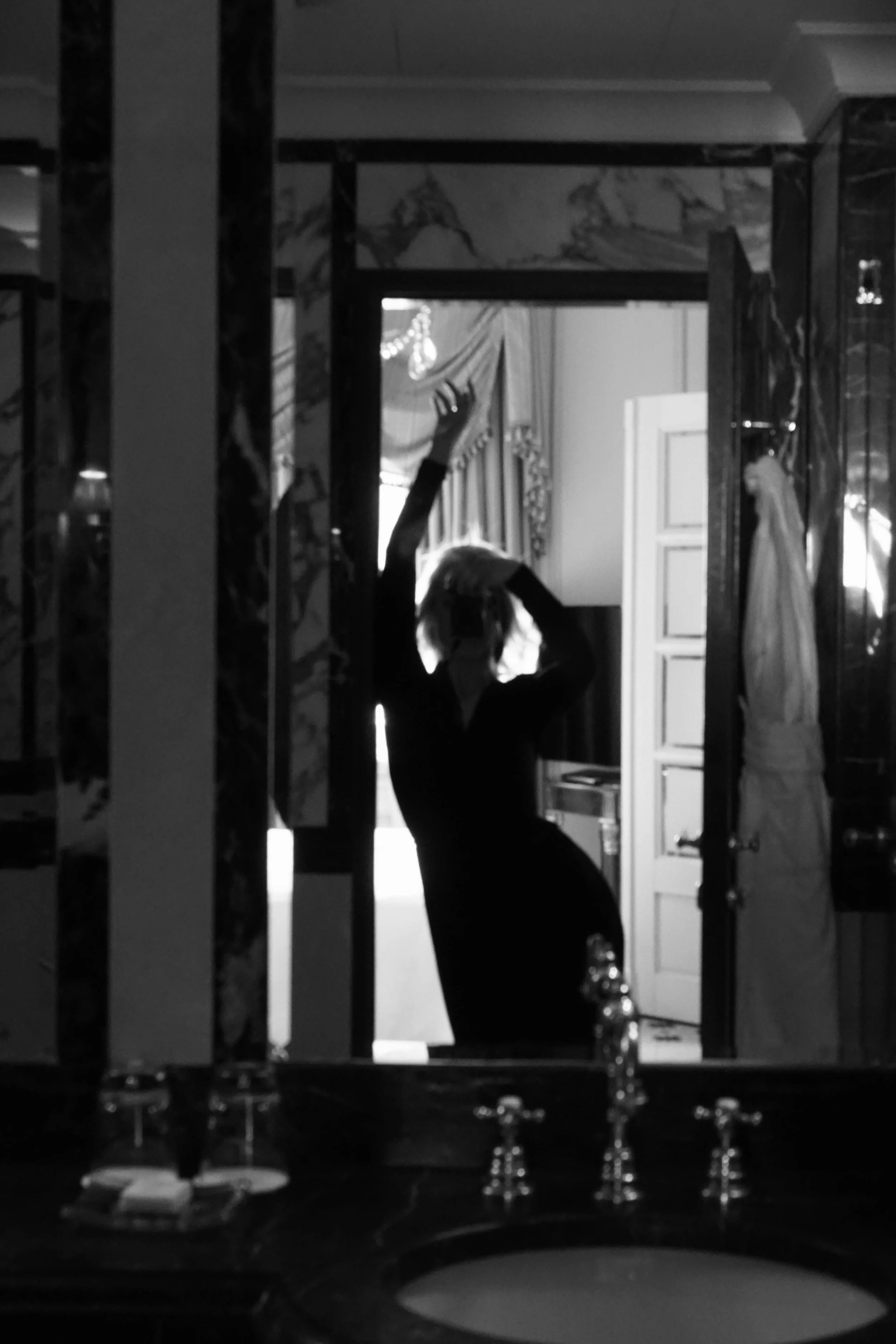 Une femme en robe noire, prenant un selfie dans un miroir, dans une pièce décorée élégamment, avec un éclairage naturel en arrière-plan.