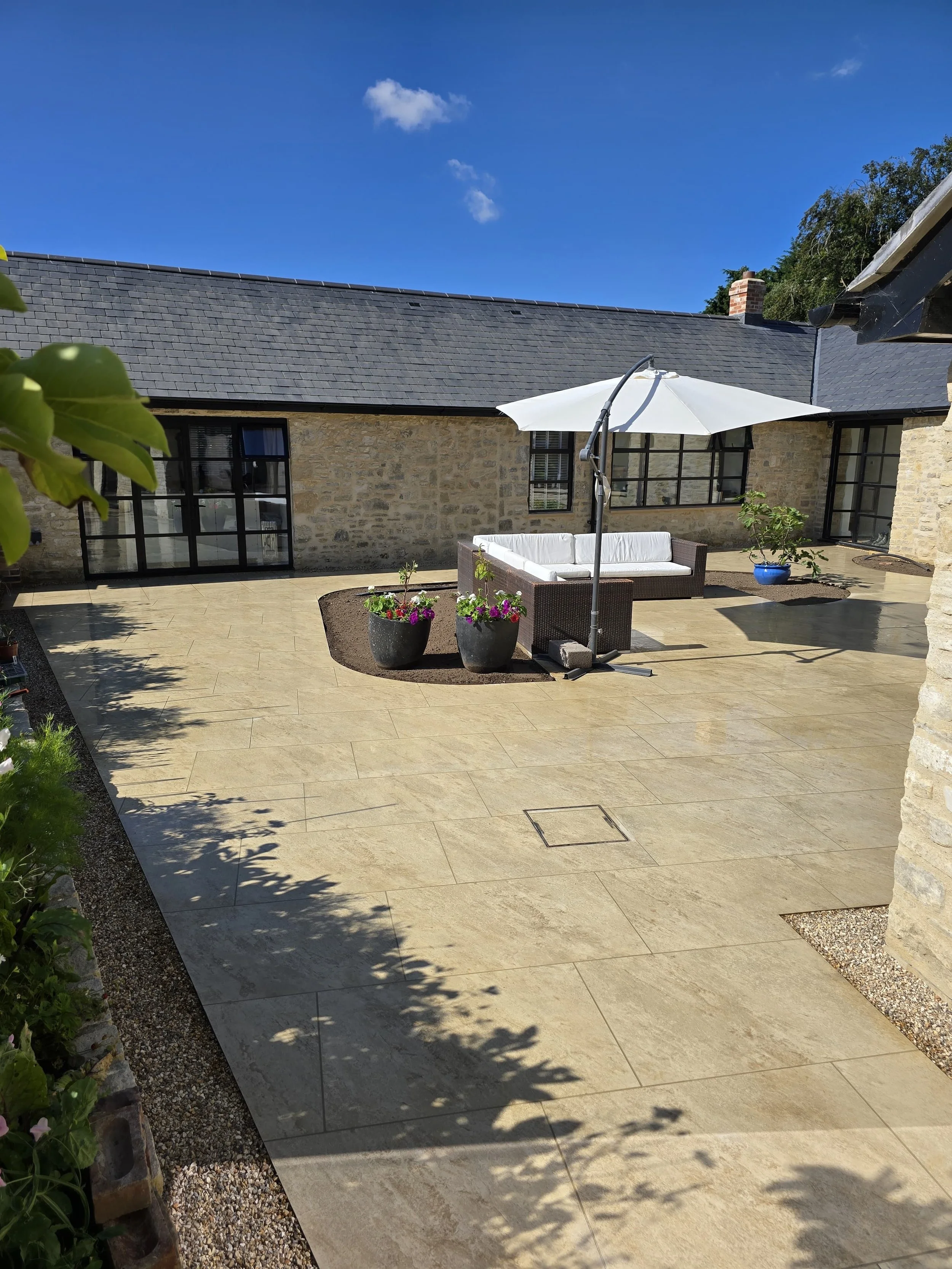 Large outdoor patio area with stone tile flooring, a white umbrella shading a wicker sofa, potted plants with colorful flowers, and a cloudy blue sky overhead.