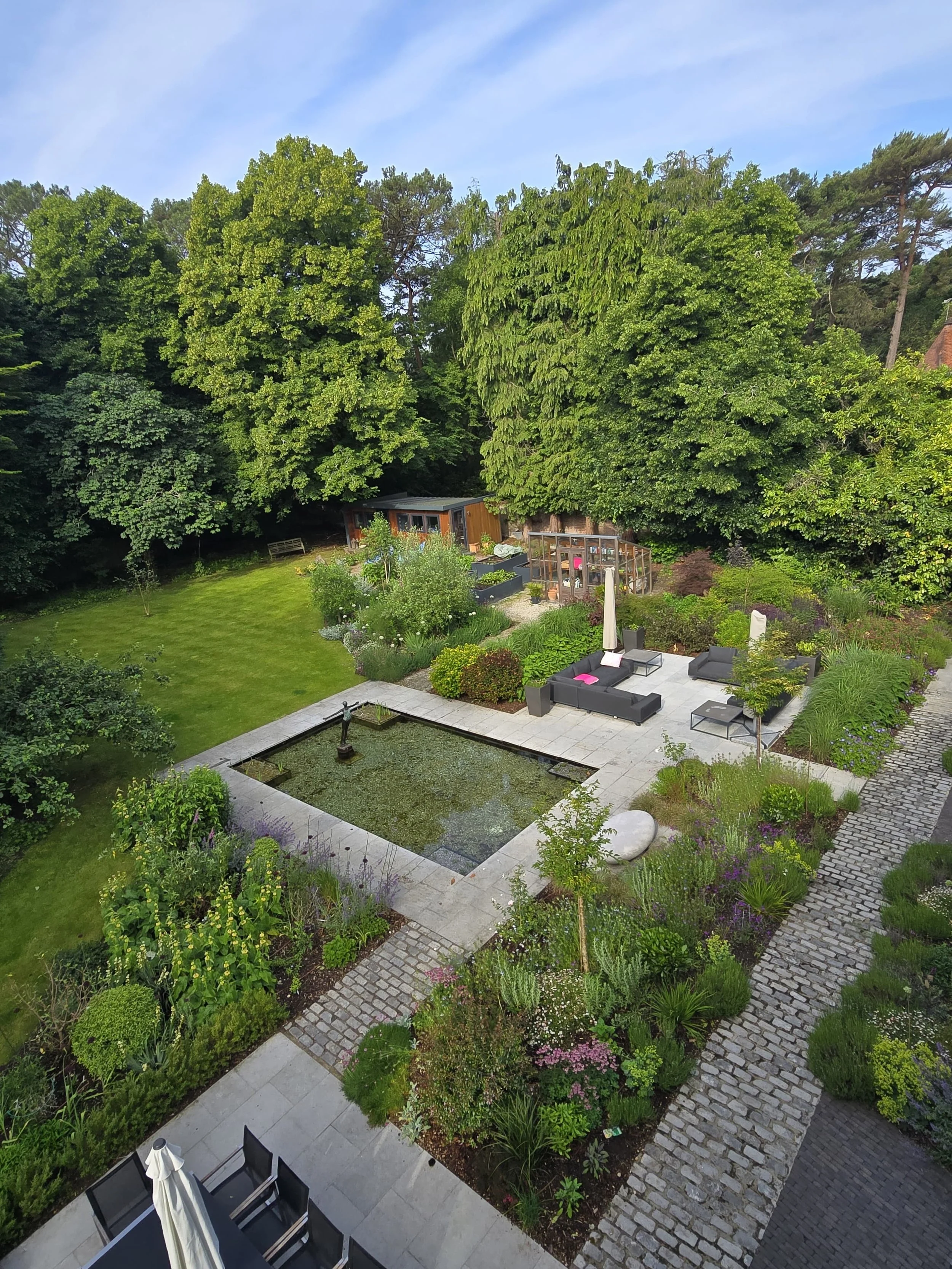 An aerial view of a well-maintained backyard garden with a small pond, patio seating area with black furniture and pink cushions, lush green lawn, garden beds with flowers and shrubs, a greenhouse, and large trees in the background.