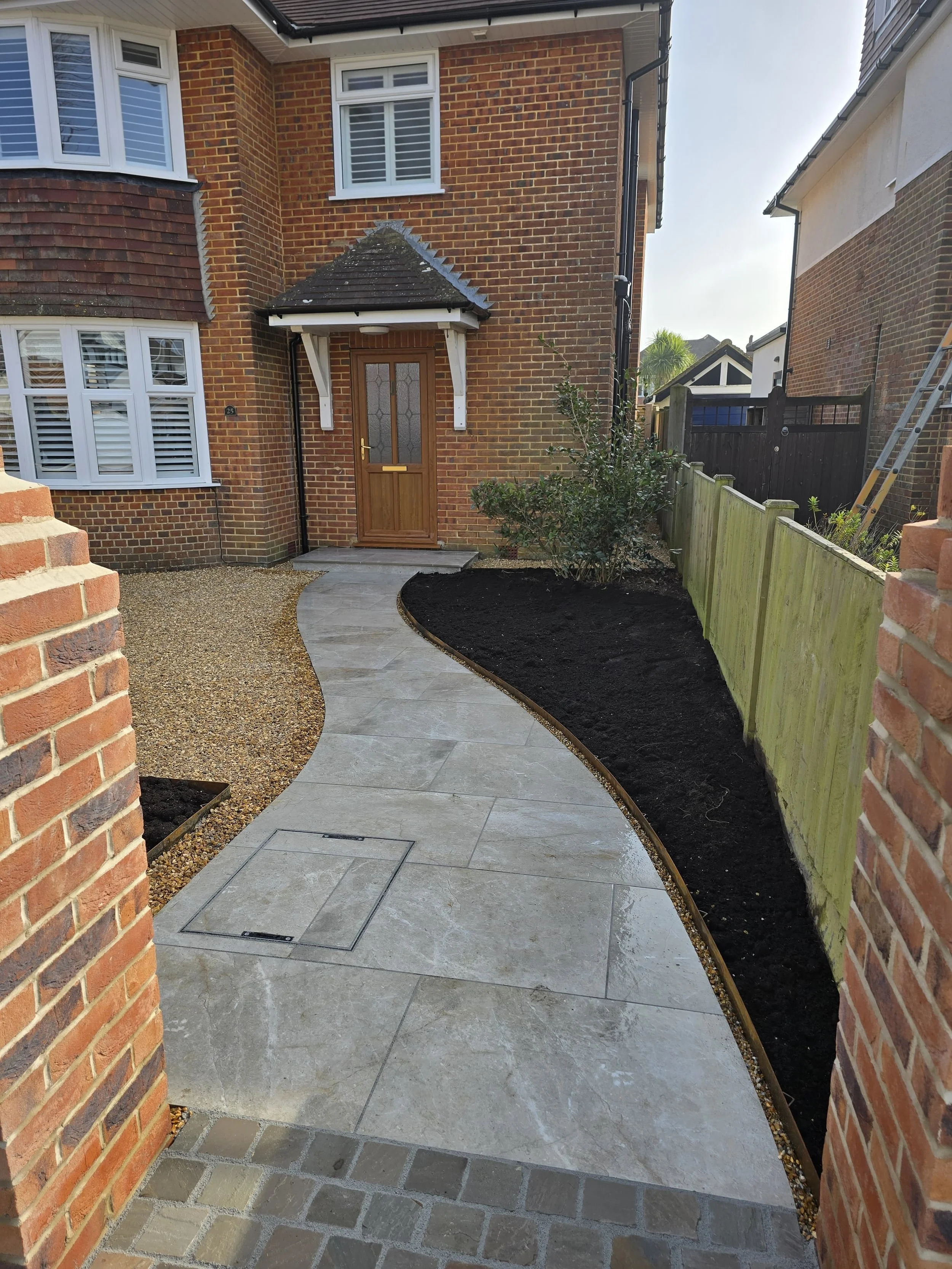 Newly paved curved walkway leading to front door of brick house, with garden bed and gravel on sides, and a wooden fence on the right.