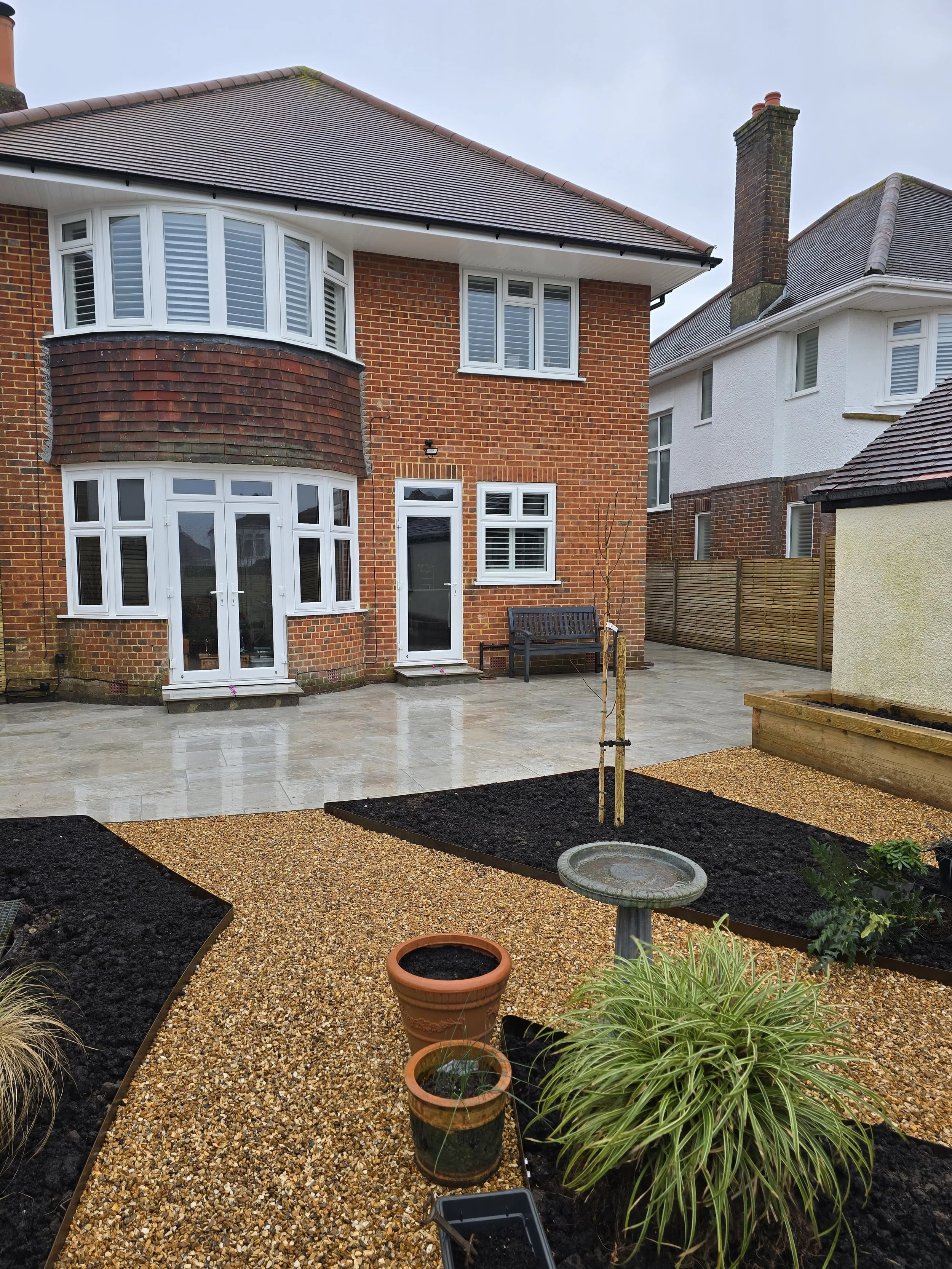 Backyard garden with newly laid gravel pathway, potted plants, a birdbath, and a young tree, with a brick house and a white house in the background.