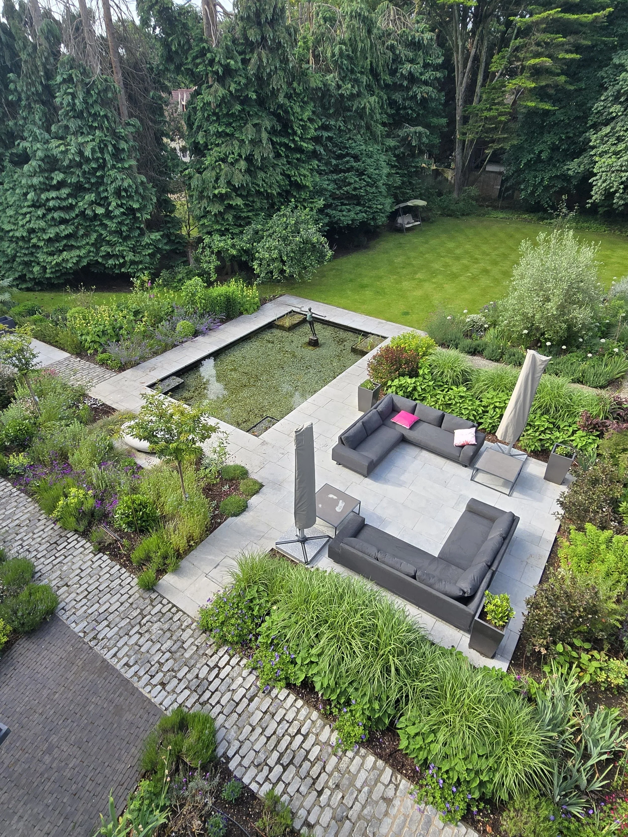 A backyard garden with a small pond, outdoor seating area with black sofas and umbrellas, lush green plants, trees, and a well-maintained lawn.