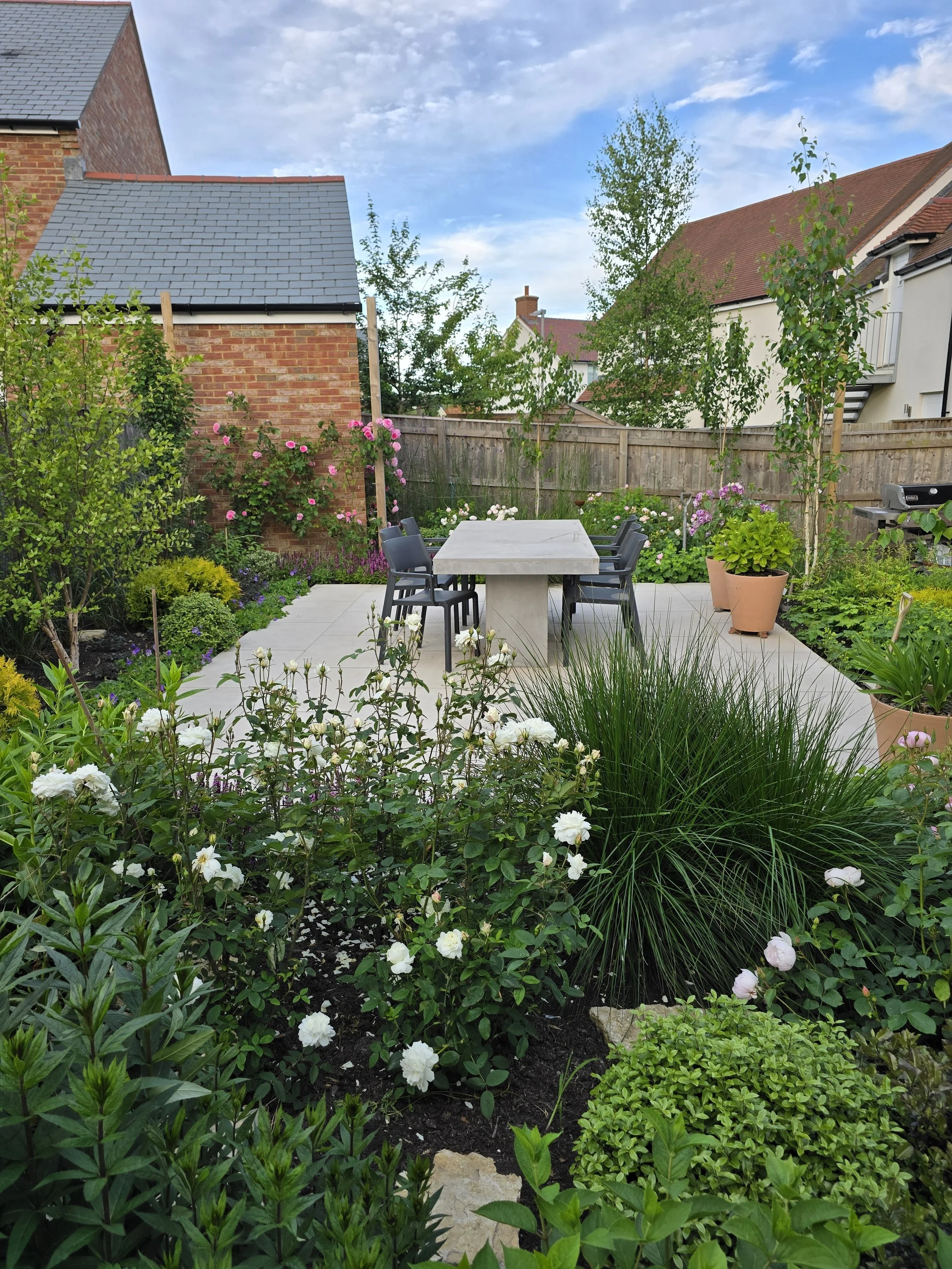 A backyard garden patio with a concrete table and six black chairs, surrounded by lush green plants and flowering bushes, with a wooden fence and neighboring houses in the background.