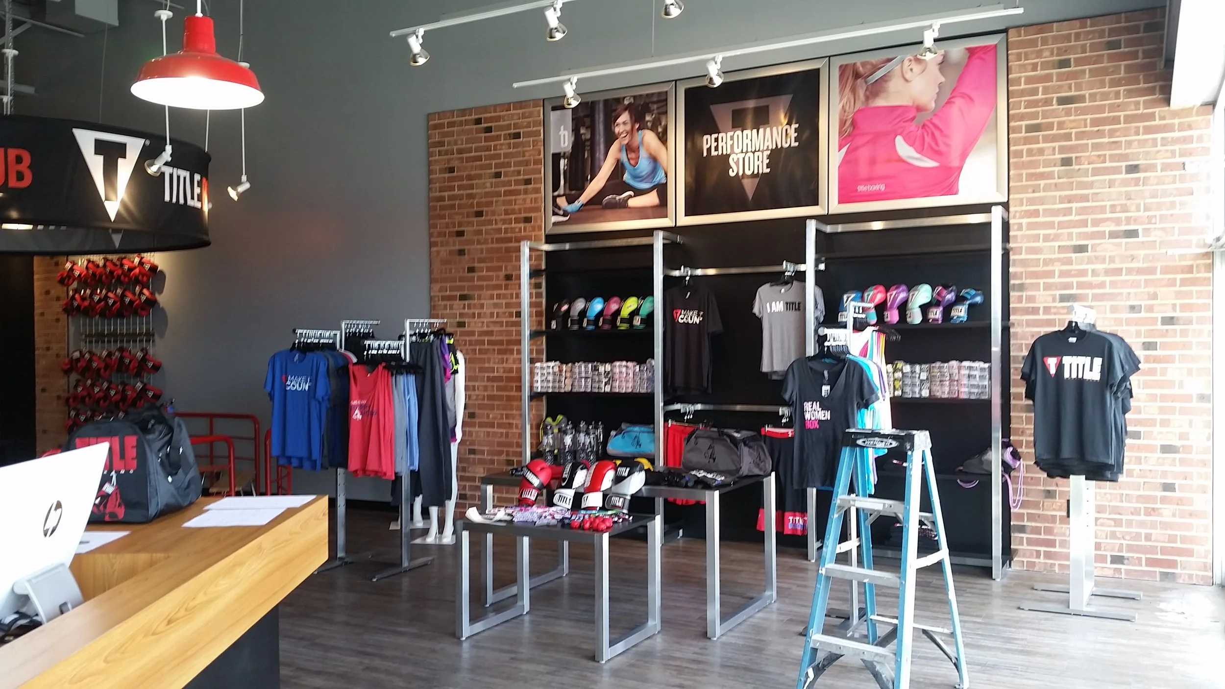 Inside a sporting goods store with workout apparel, shoes, and equipment on display, including shelves of colorful shoes and T-shirts, plastic gloves, and boxing gloves, with promotional posters on the brick wall.