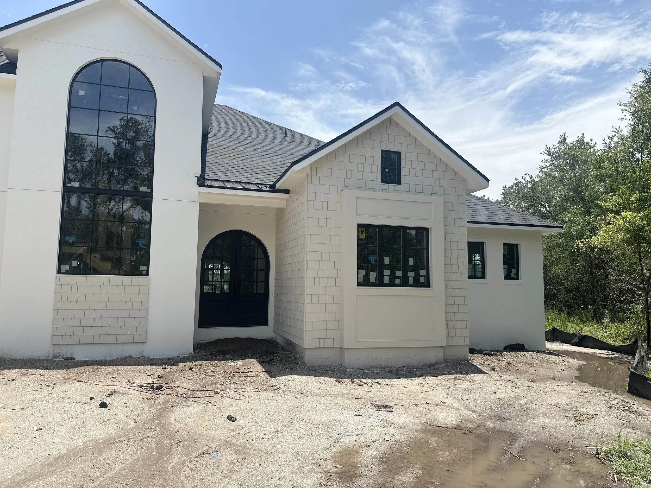 Newly constructed white house with large windows and black doors, on a dirt lot with some greenery and trees in the background.