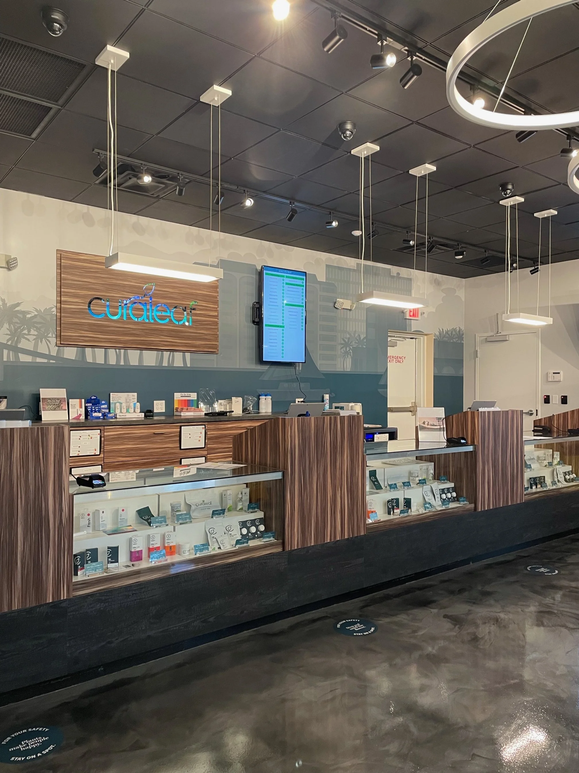 Interior of a medical dispensary with a modern wood and glass display counter, digital menu screen, and a wall sign that reads 'Curaleaf.'
