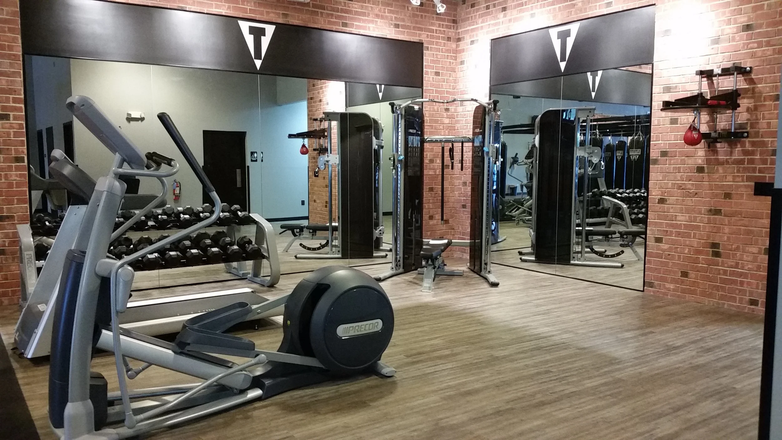 Gym with exercise equipment including a treadmill, elliptical, weight rack, and cable machine, reflected in large mirrors on brick wall.