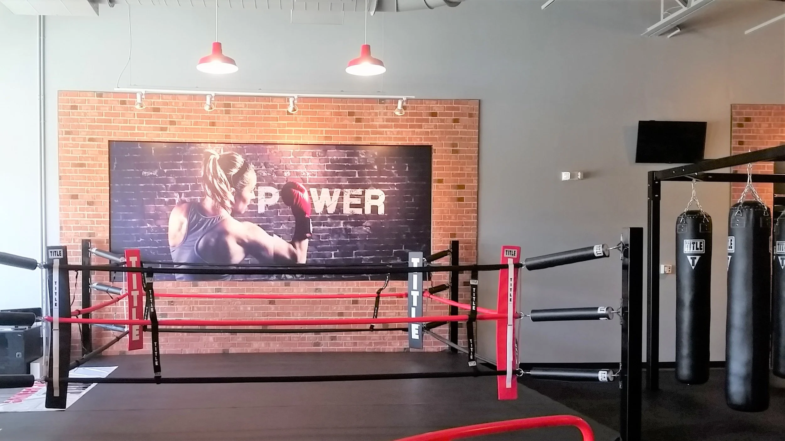 Empty boxing ring with red and black ropes, punching bags, and a large wall poster of a woman boxing with the word 'POWER' in the background.