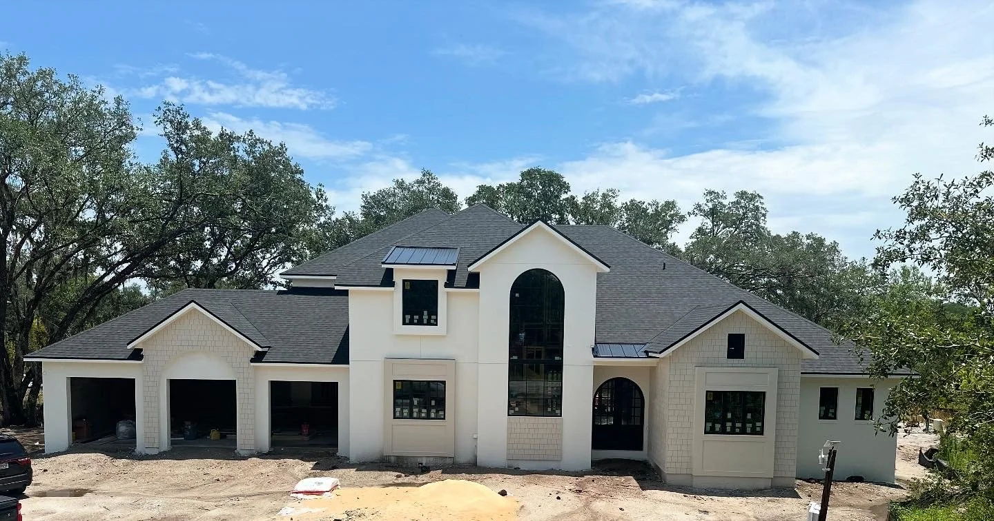 Newly built two-story house under construction with a dark gray roof, large arched window in the center, and multiple smaller windows. The house has a white exterior and is surrounded by trees and construction debris.