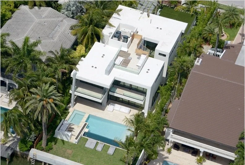 Aerial view of a modern white house with a rooftop terrace, a backyard with a swimming pool, surrounded by tall palm trees and neighboring houses in a lush, tropical setting.