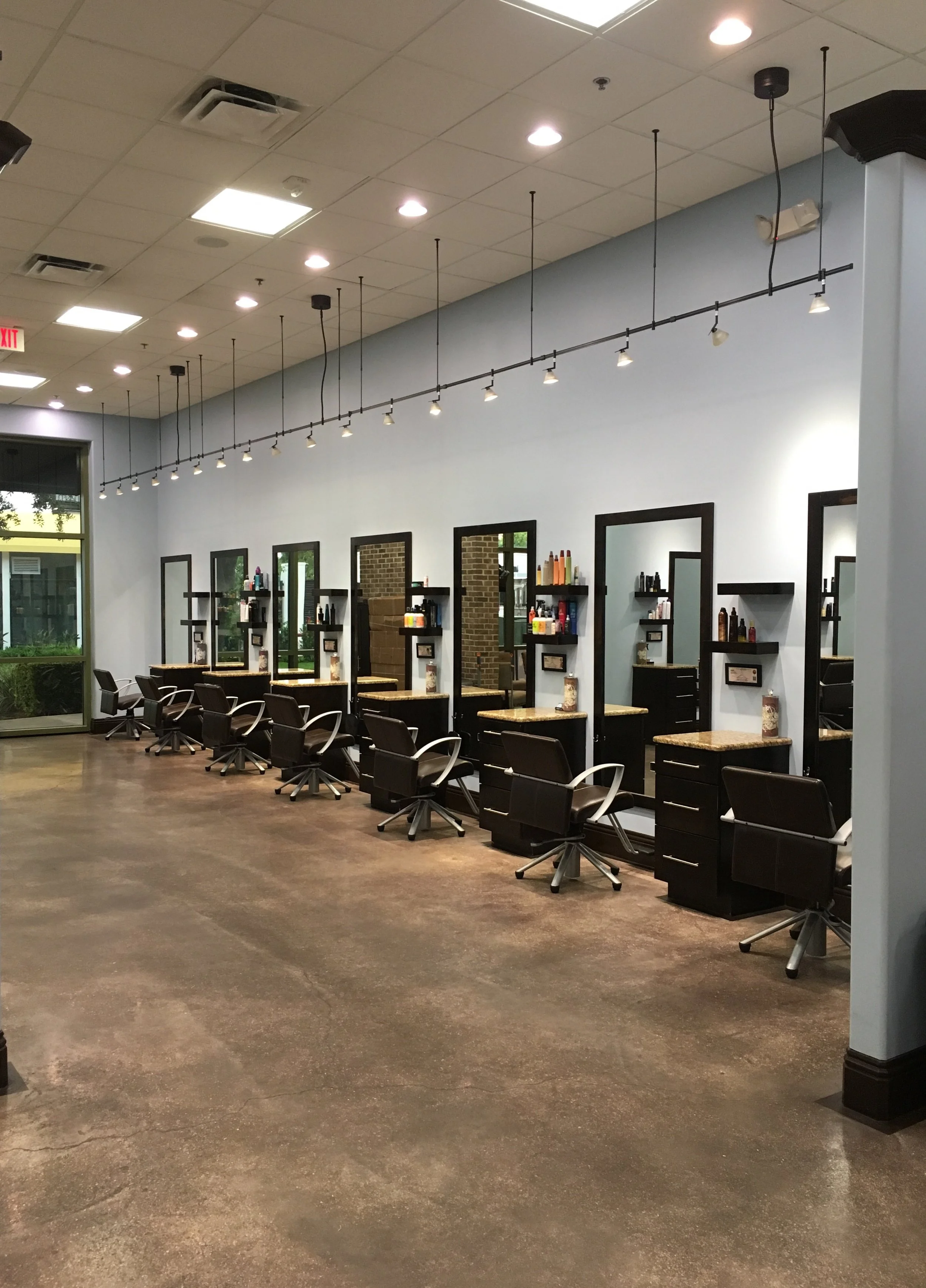 Empty hair salon with multiple styling stations, mirrors, chairs, and hair products, illuminated by ceiling lights and track lighting.